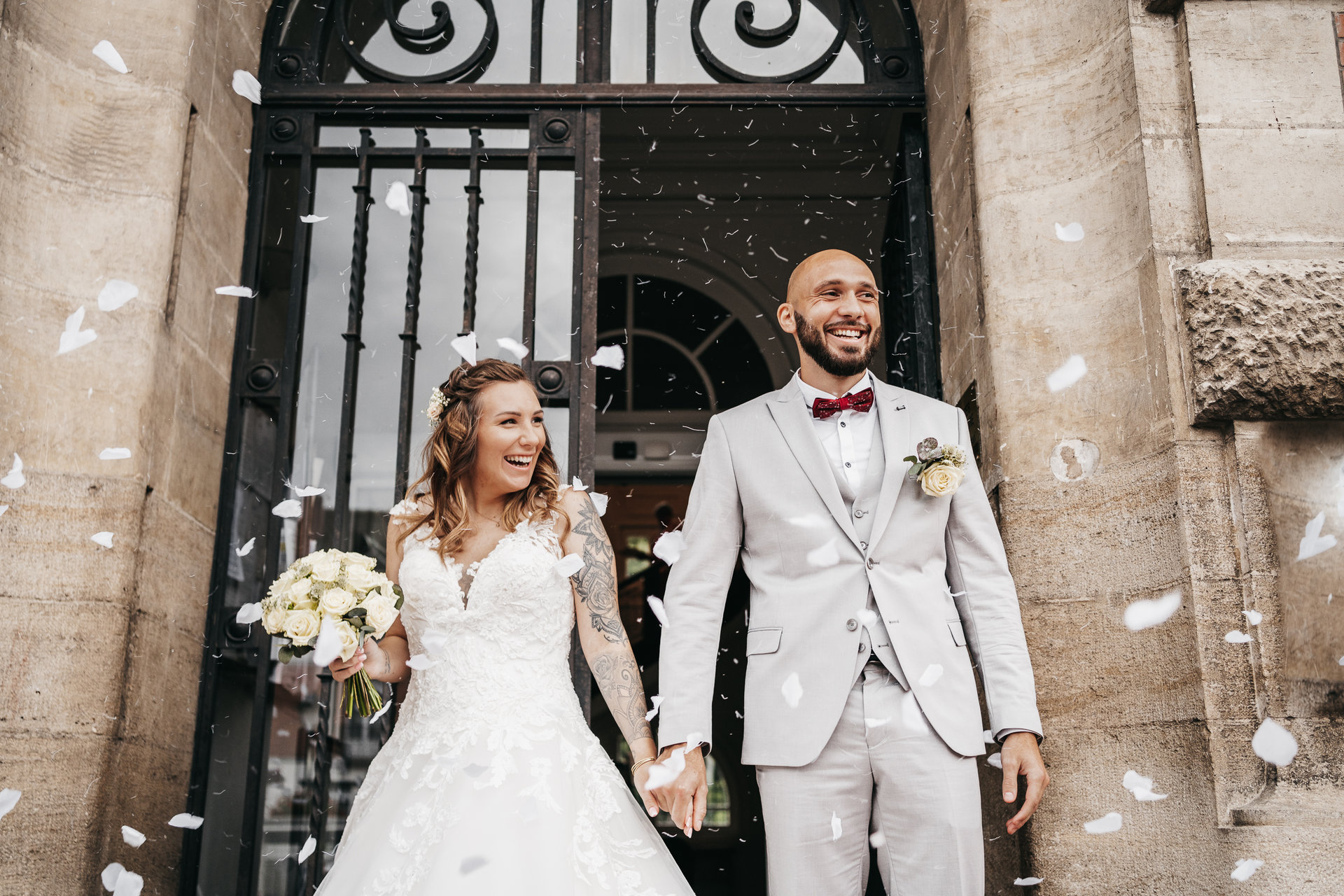 Une mariée et un marié sortent d'un immeuble avec des confettis lancés sur eux.