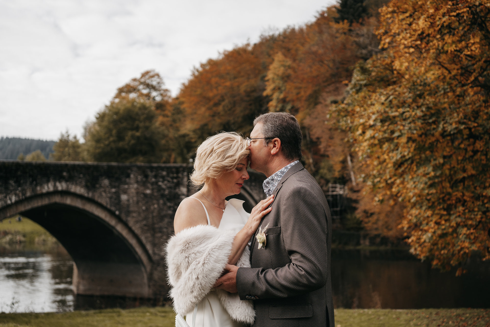 Une mariée et un marié s'embrassant devant un pont en automne.