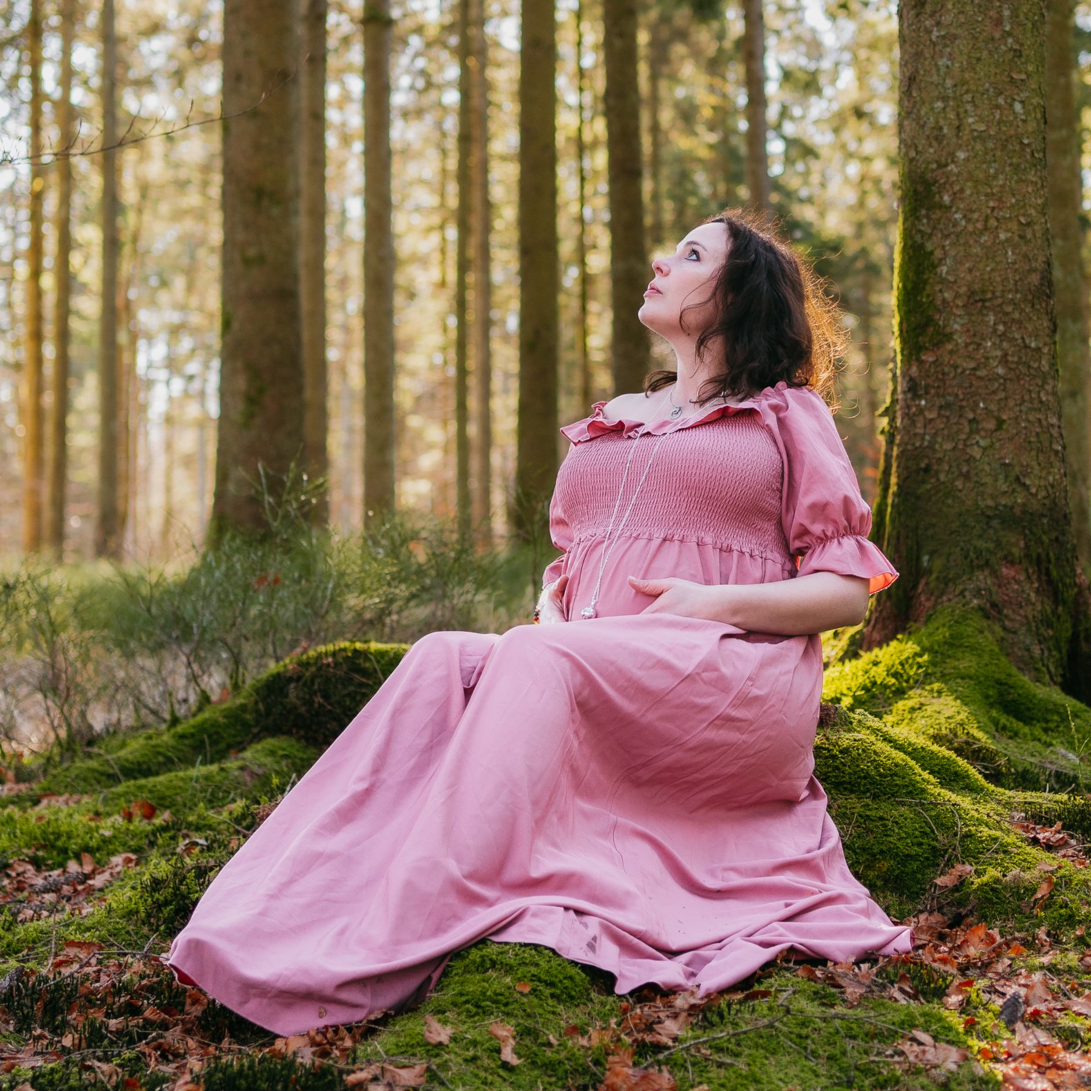 Une femme enceinte vêtue d'une robe rose assise sur de la mousse dans une forêt.