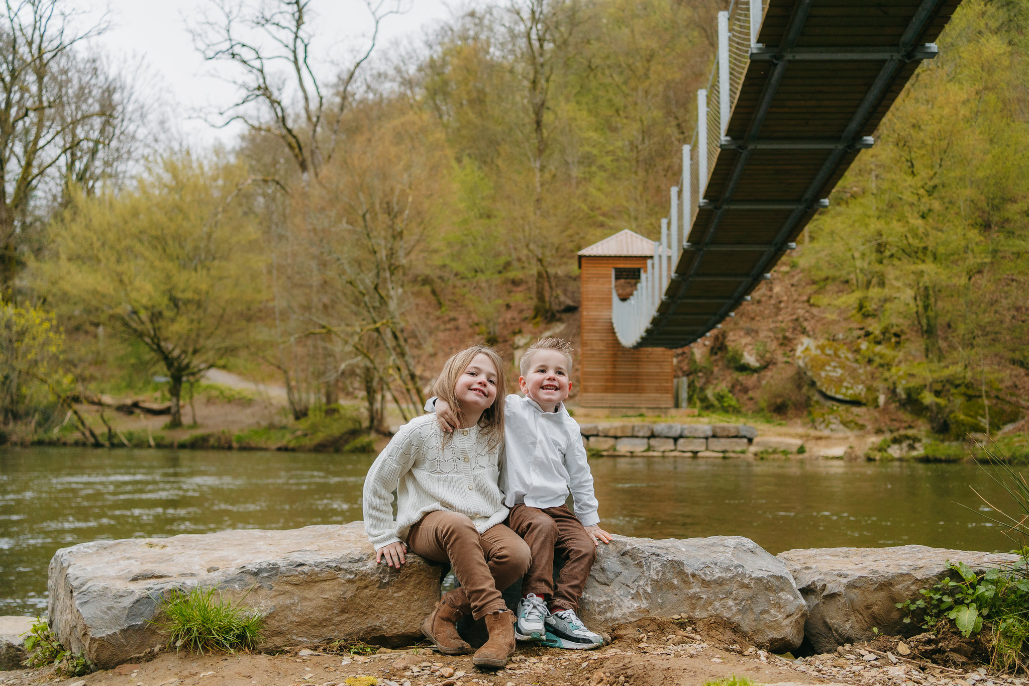 Deux enfants assis sur des rochers près d’un pont suspendu.