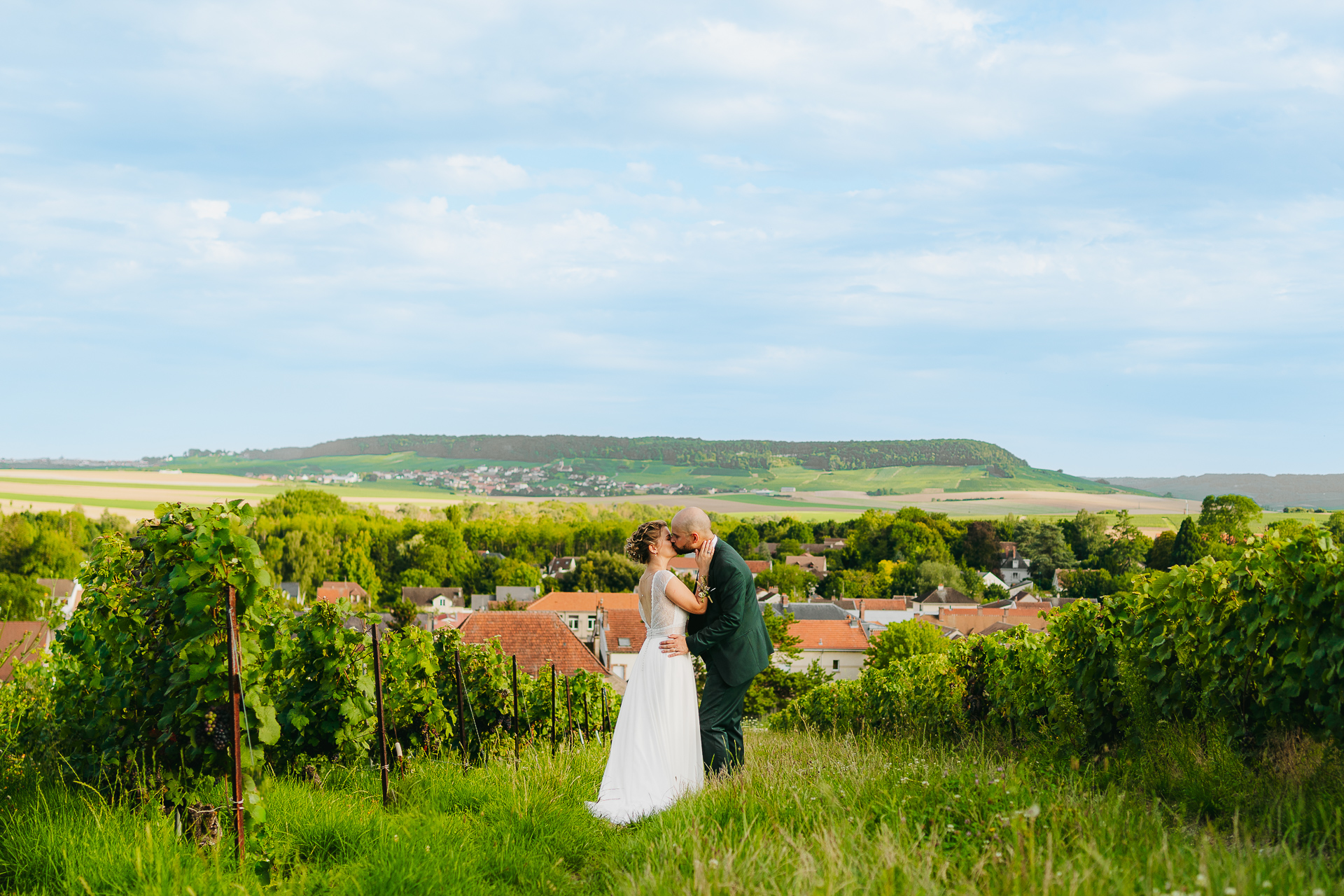 Couple enlacé dans un vignoble avec une vue panoramique sur les collines et un village en arrière-plan.