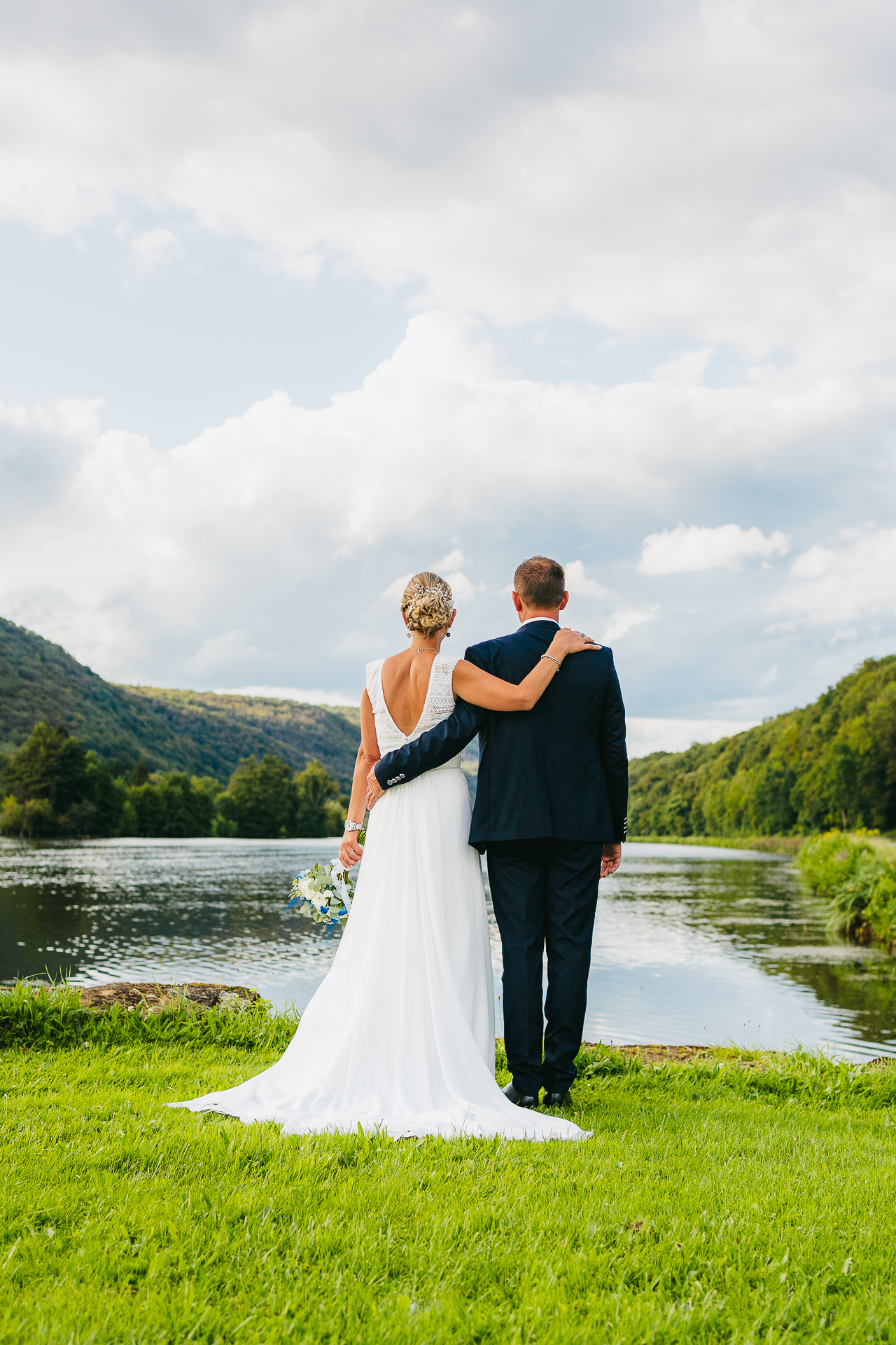 Un couple de jeunes mariés debout au bord d’un lac, regardant le paysage, dos à la caméra.