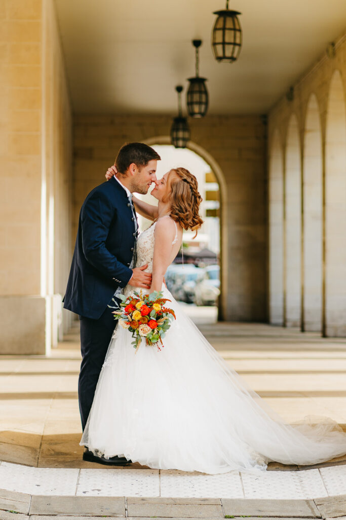 Des mariés partageant un baiser tout en tenant un bouquet dans une arcade avec des lanternes suspendues, capturés par le photographe mariage Reims.