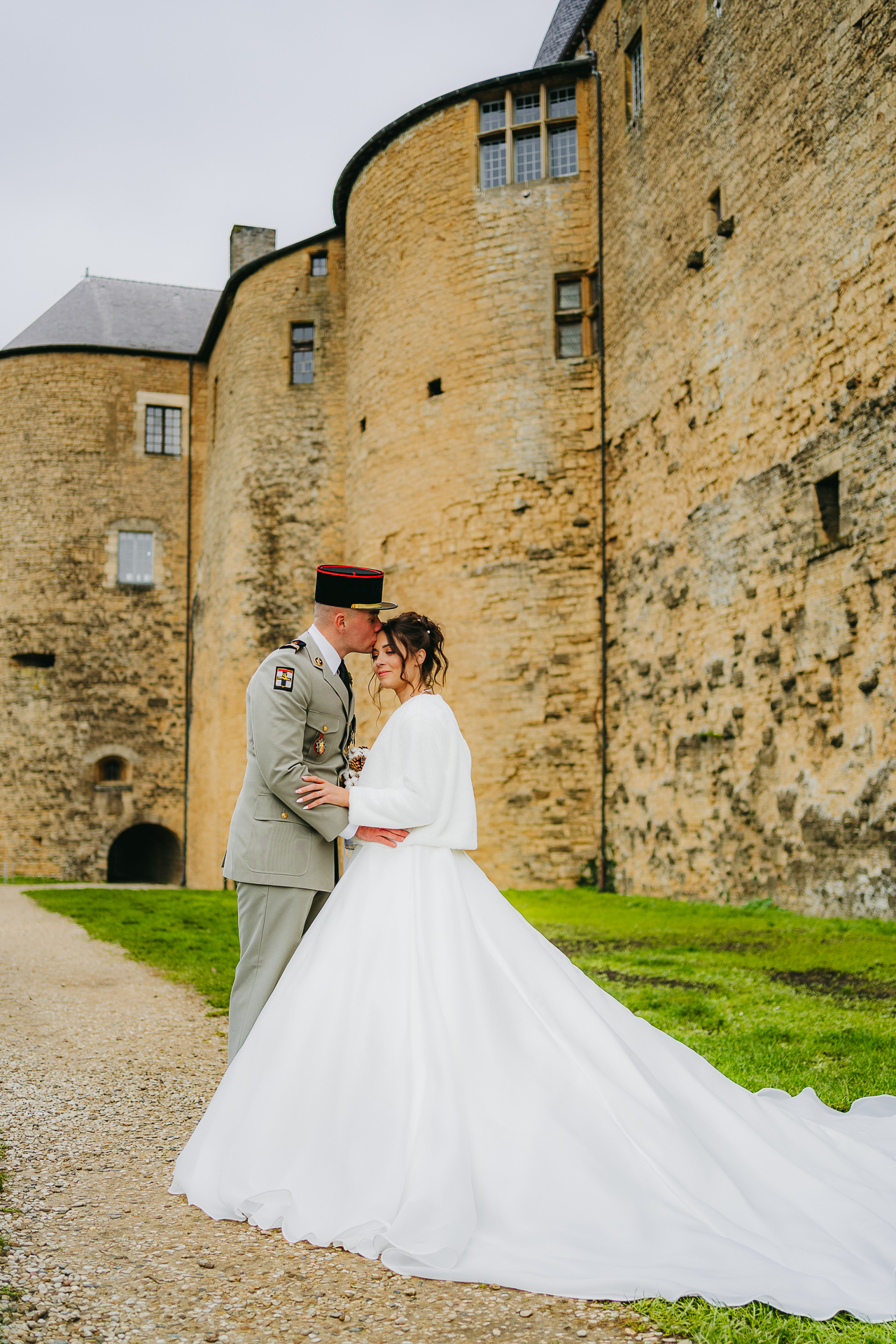 Un individu en uniforme et une personne en robe de mariée blanche partagent un moment intime à côté d’un ancien château de pierre.