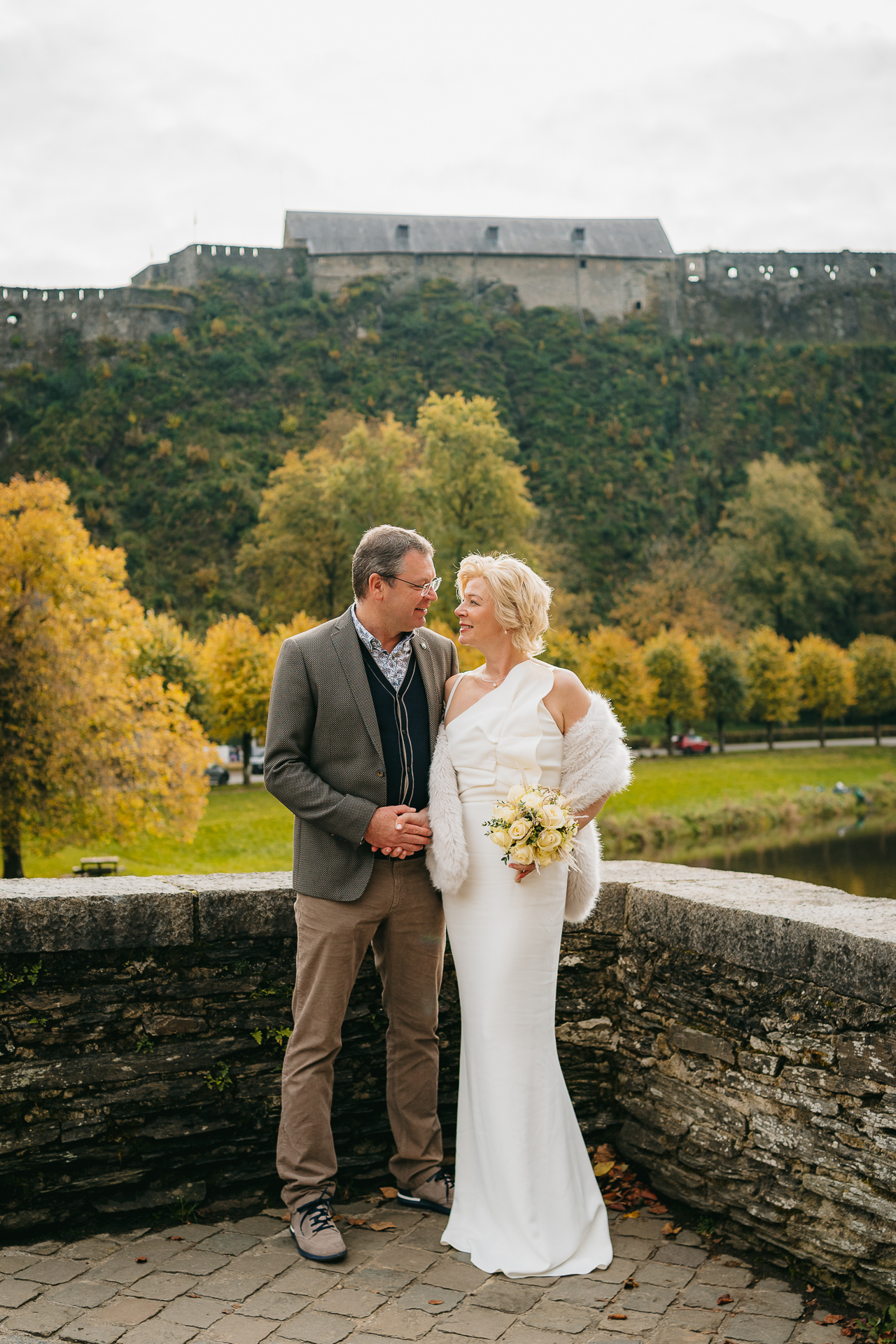 Un couple en tenue de mariage partageant un moment affectueux avec une forteresse historique et un feuillage d'automne en arrière-plan.