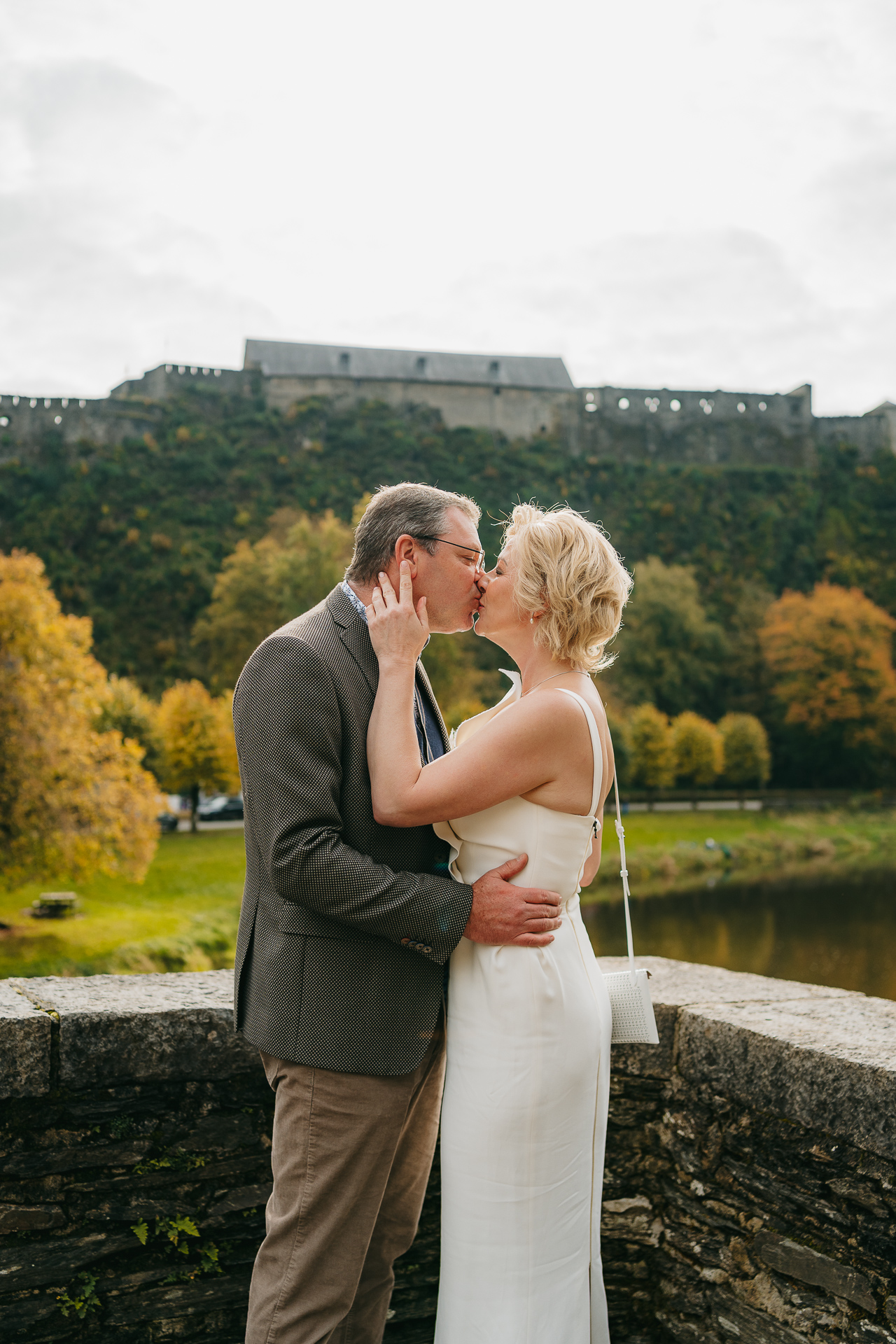 Un couple partageant un baiser sur un pont avec un château historique et des arbres automnaux en arrière-plan.