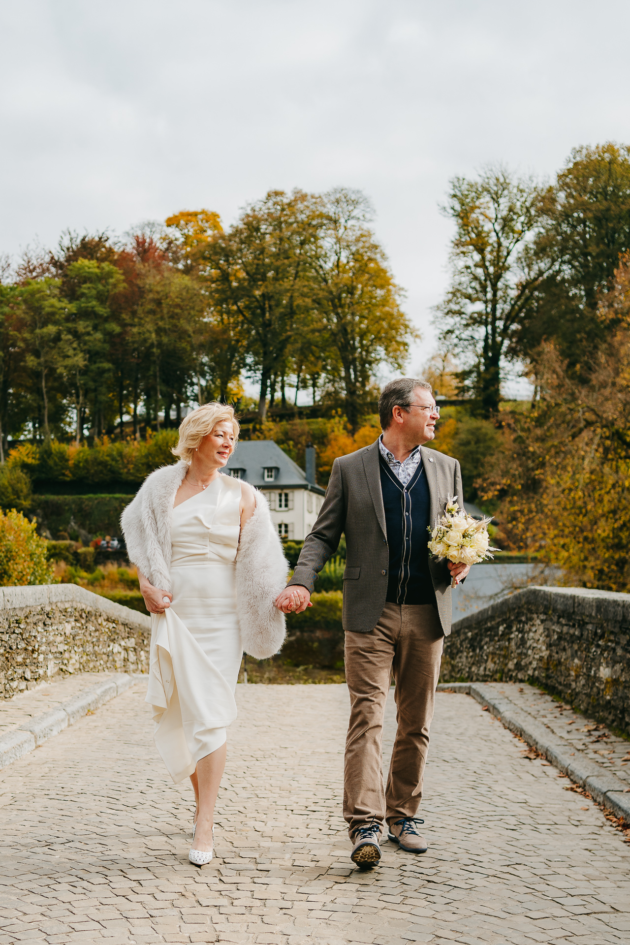 Un couple en tenue de mariage se tenant la main en traversant un pont de pierre avec des feuilles d'automne en arrière-plan.