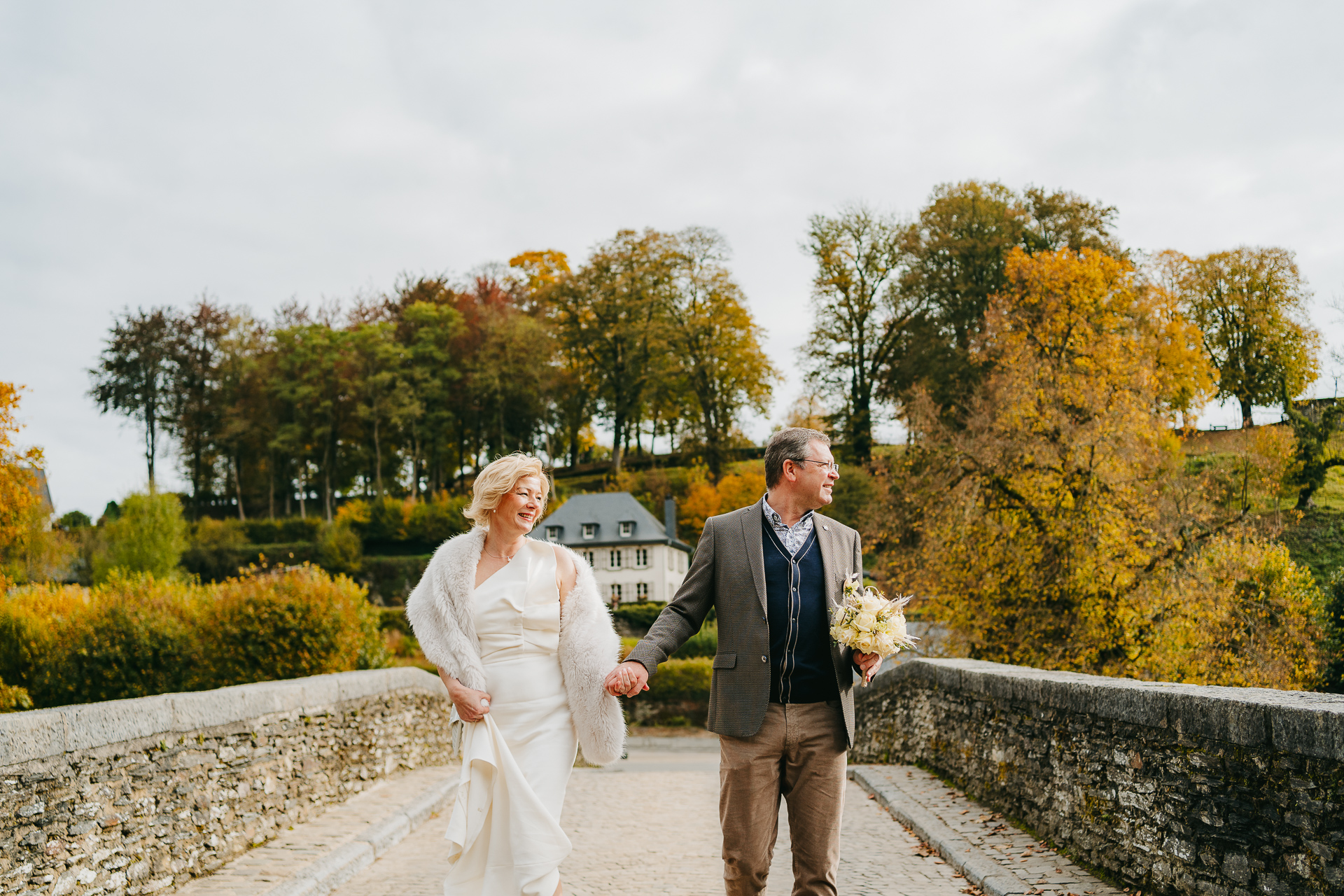 Un couple se tenant la main et traversant un vieux pont de pierre avec des arbres d'automne en arrière-plan.