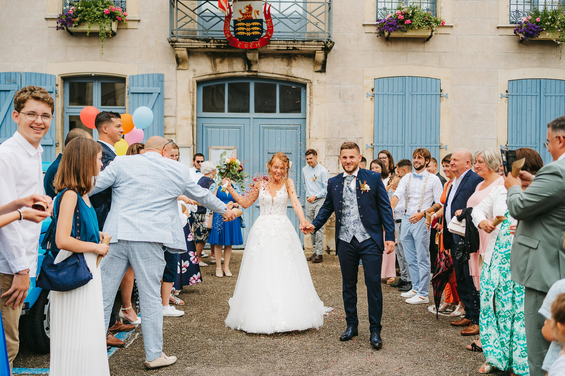 Des mariés se tiennent la main, souriants, alors qu'ils traversent une foule d'invités devant un bâtiment aux volets bleus et aux bacs à fleurs. Dans cette magnifique photo de reportage de mariage, les invités applaudissent et tiennent des ballons.