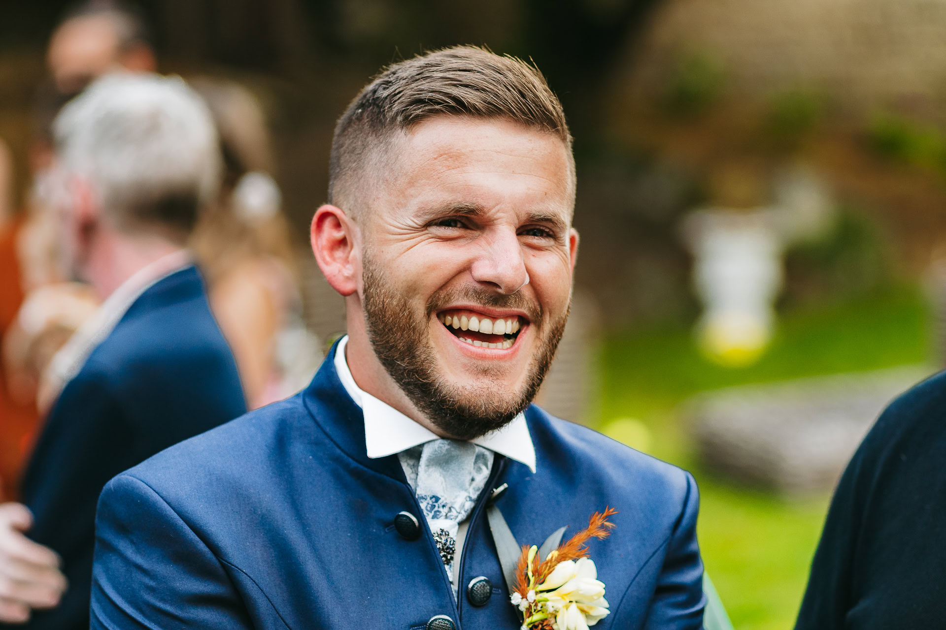 Un homme aux cheveux courts et à la barbe sourit largement en portant un costume bleu avec une boutonnière sur cette photo de reportage de mariage. L’arrière-plan est flou avec des personnages et de la verdure, capturant l’essence de cette joyeuse occasion.