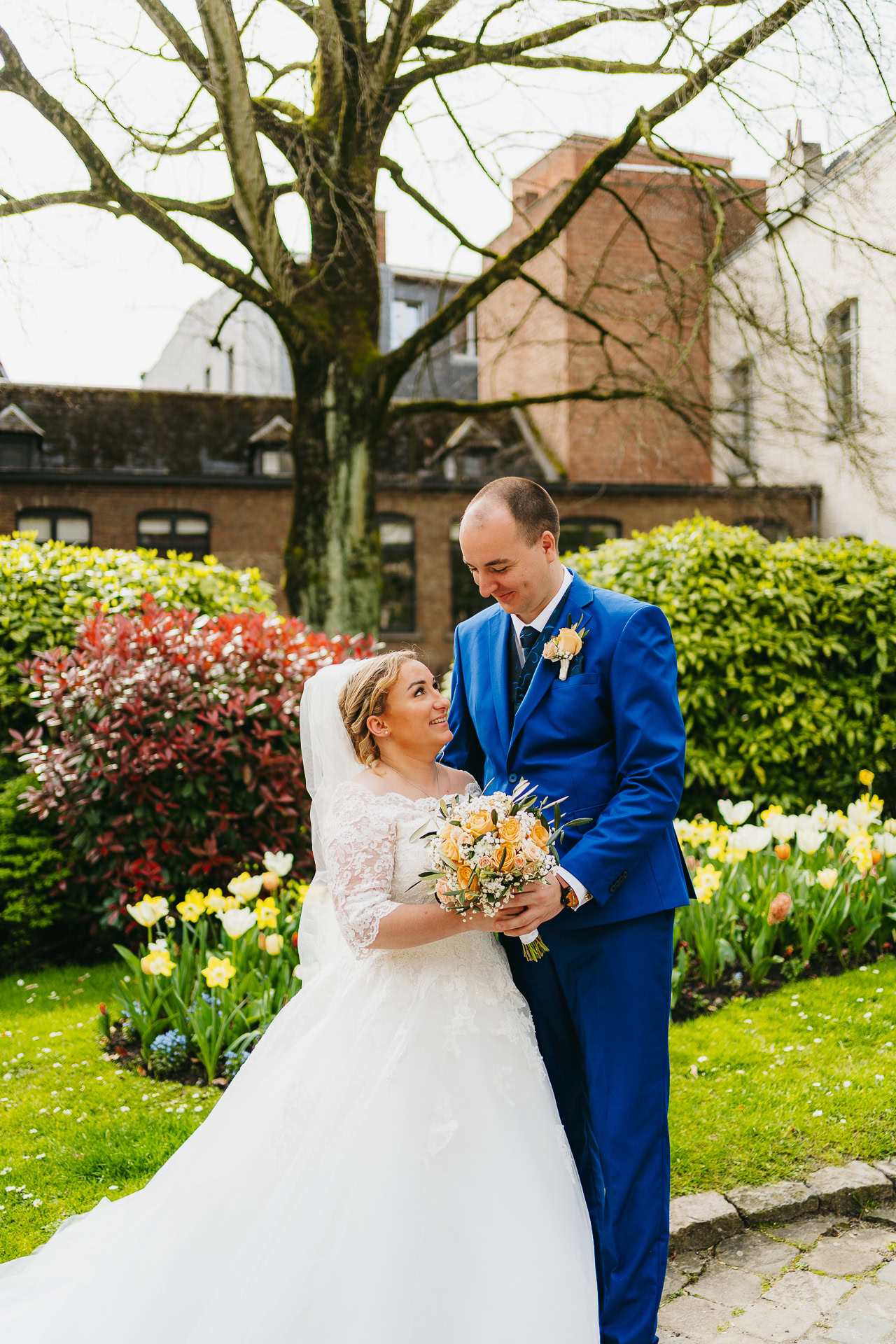 Une mariée en robe blanche et un marié en costume bleu se sourient, debout dans un jardin coloré avec des fleurs épanouies.