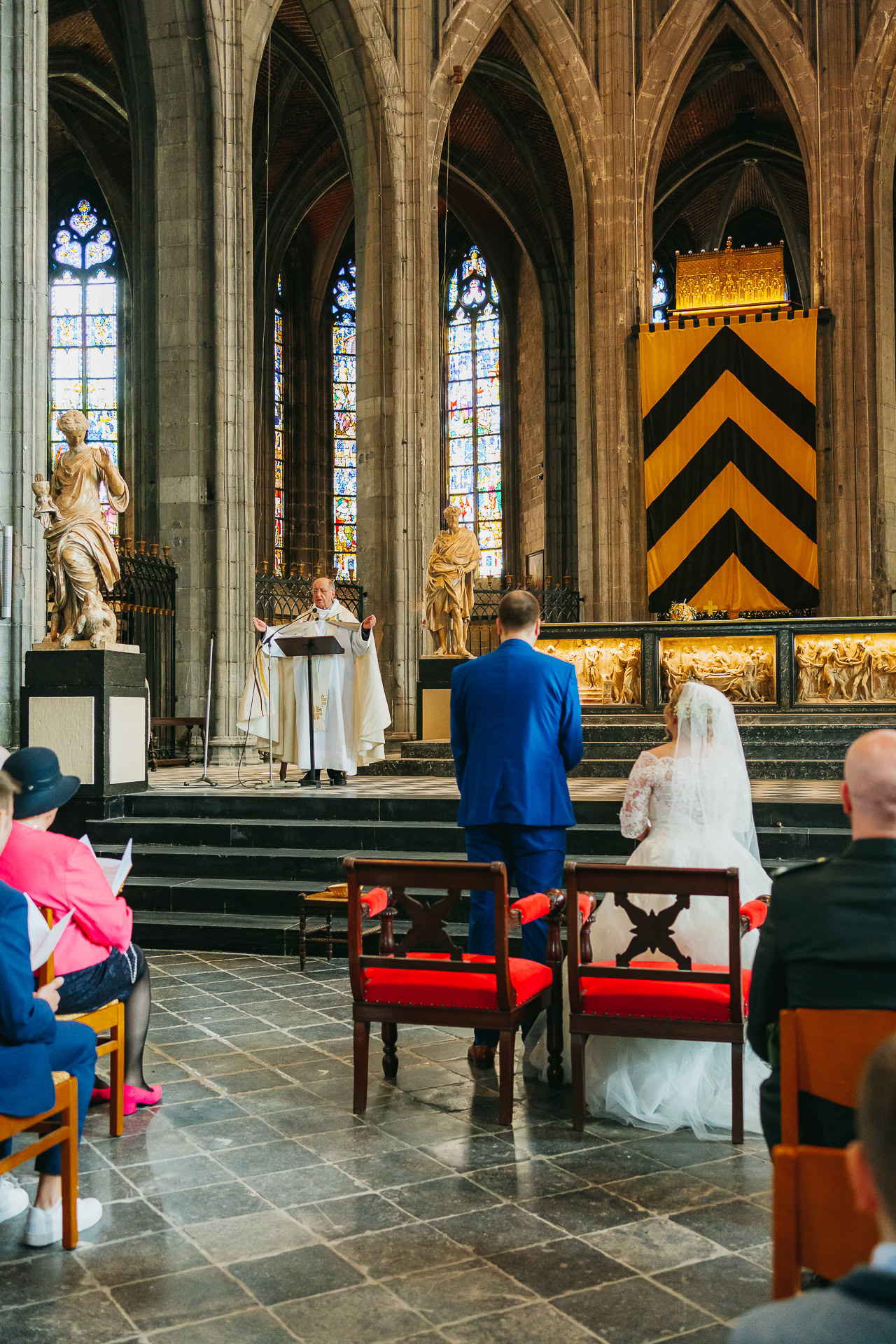 Des mariés se tiennent devant un prêtre dans une cathédrale lors d'une cérémonie de mariage, avec des invités assis autour d'eux.