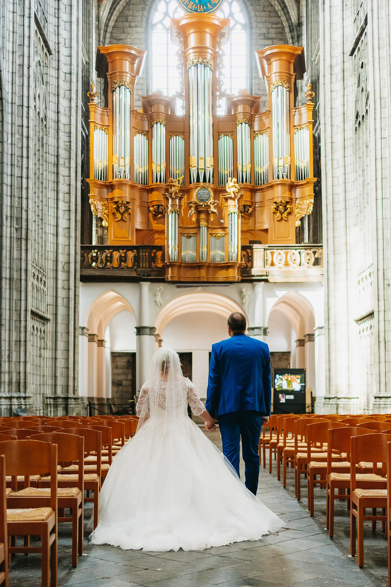 La mariée en robe blanche et le marié en costume bleu marchent main dans la main dans l'allée d'une cathédrale avec un grand orgue en arrière-plan.