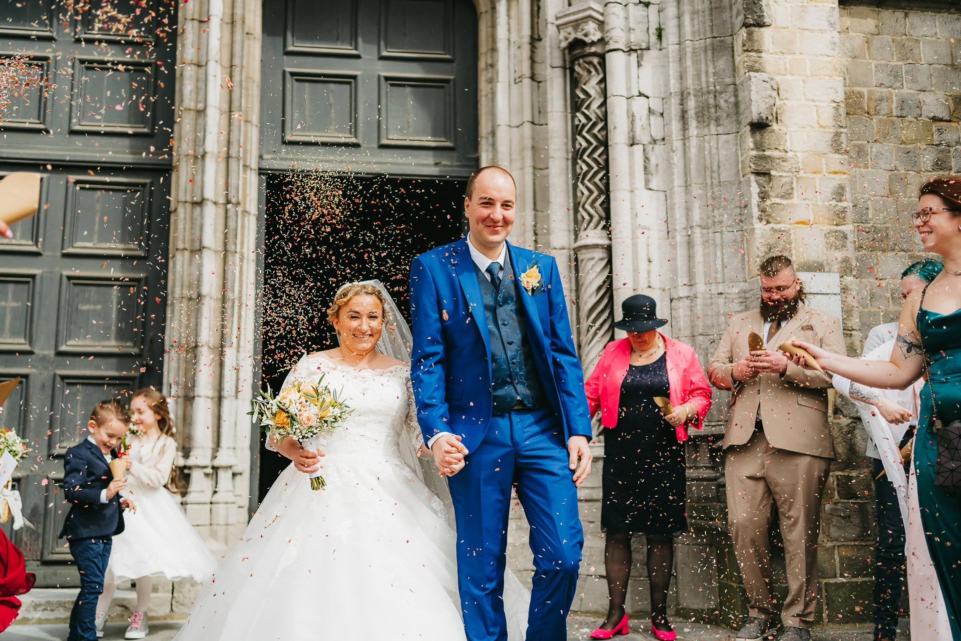 Un couple de jeunes mariés sourit en marchant sous une pluie de confettis, entouré d’invités devant une église.