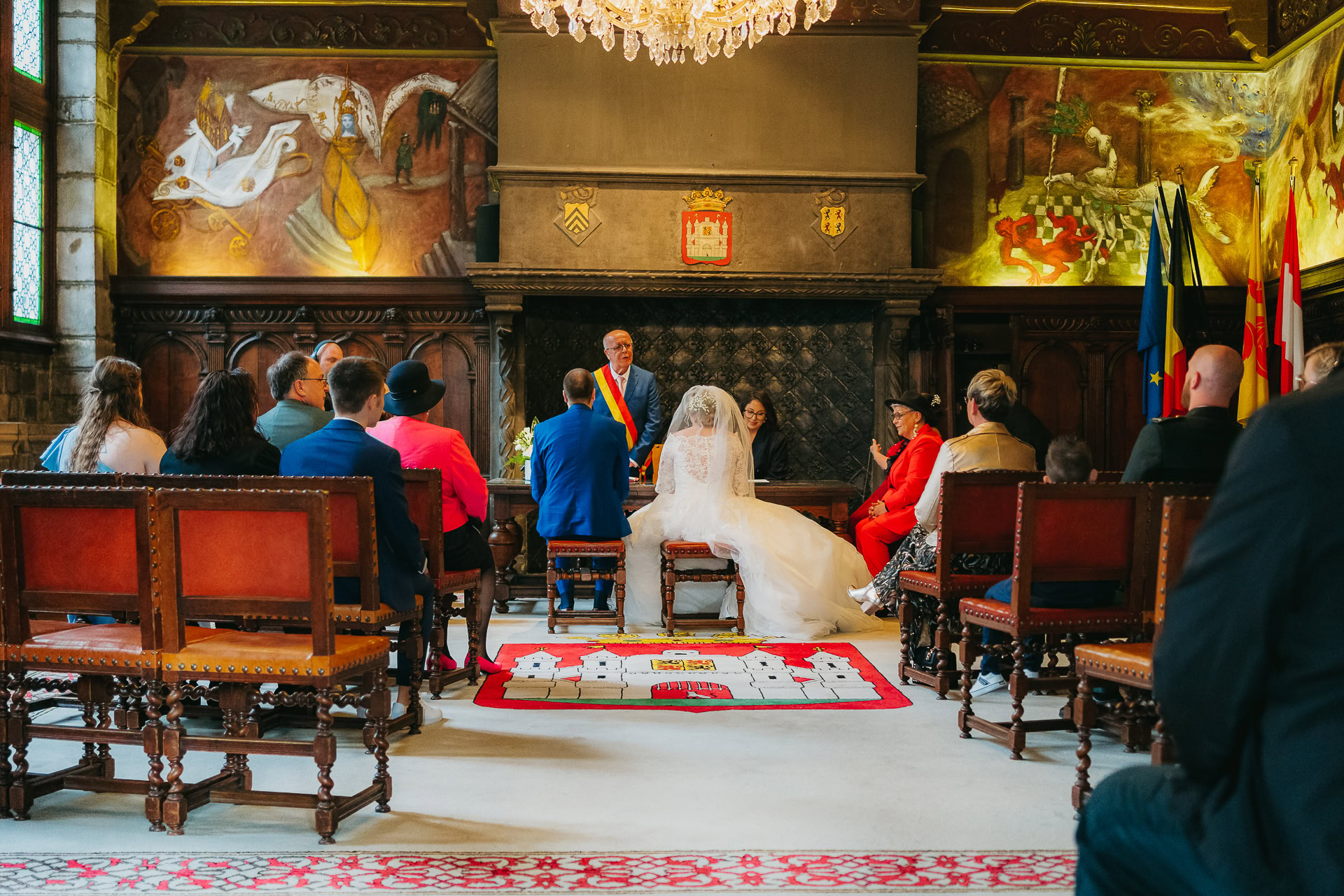 Cérémonie de mariage dans une grande salle ornée de peintures murales et d'armoiries richement détaillées, mettant en vedette les mariés assis face à un orateur, entourés d'invités.