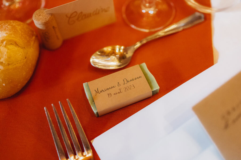Un couvert avec un rouleau, une fourchette, une cuillère, une étiquette nominative en papier et un petit objet emballé étiqueté « Marianne & Donovan 19 août 2023 » sur une nappe rouge présente des savons personnalisés mariage.
