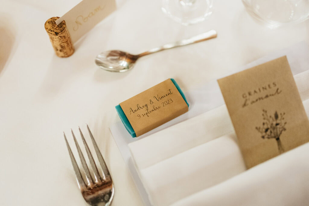 Gros plan d'une table de mariage avec une carte de visite, une cuillère, une fourchette, une serviette avec un emballage étiqueté « Audrey & Vincent 9 septembre 2023 », savons personnalisés mariage et une petite carte intitulée « Graines d'amour ».