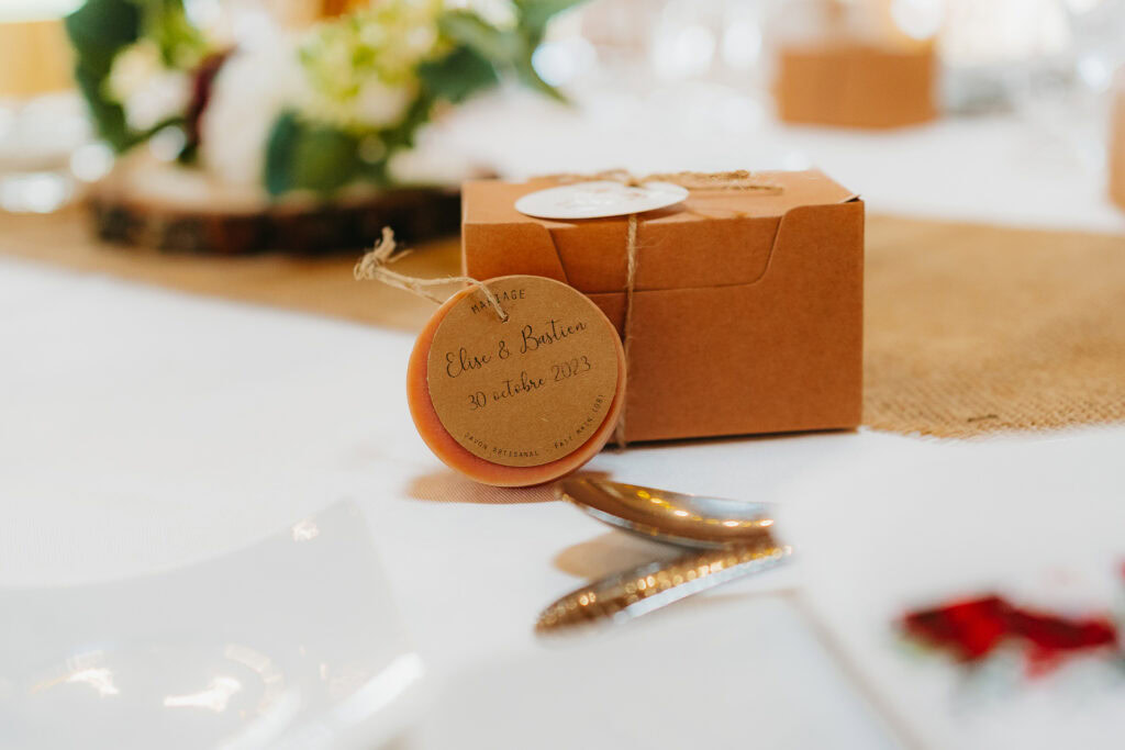 Une petite boîte en carton marron posée sur une table, nouée avec de la ficelle et une étiquette ronde sur laquelle est écrit « Ella & Braden 7 octobre 2023 ». L’arrière-plan montre un arrangement floral flou et des réglages de table, faisant allusion aux cadeaux de mariage personnalisés à l’intérieur.