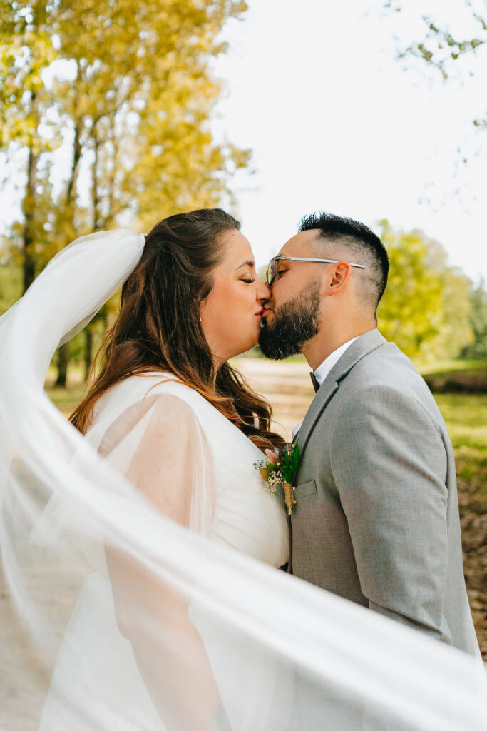 Les mariés partagent un baiser en plein air le jour de leur mariage. Le voile de la mariée flotte et elles sont entourées d'arbres aux feuilles jaunes.