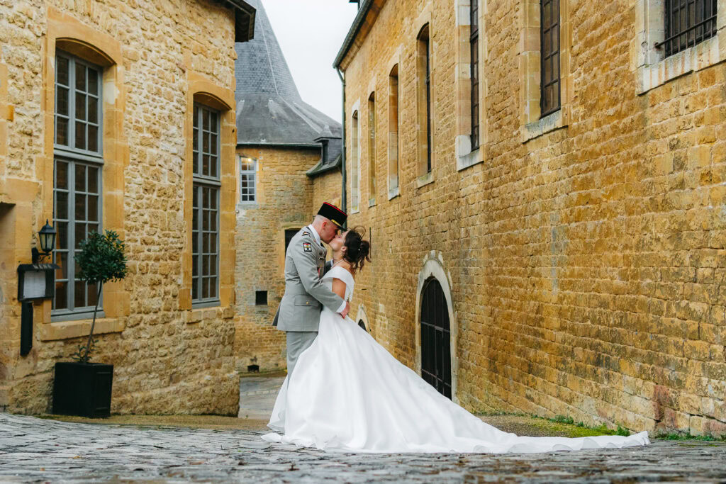 Un couple, vêtu d'un uniforme militaire et d'une robe de mariée, partage un baiser dans une étroite ruelle pavée bordée de vieux bâtiments en pierre.