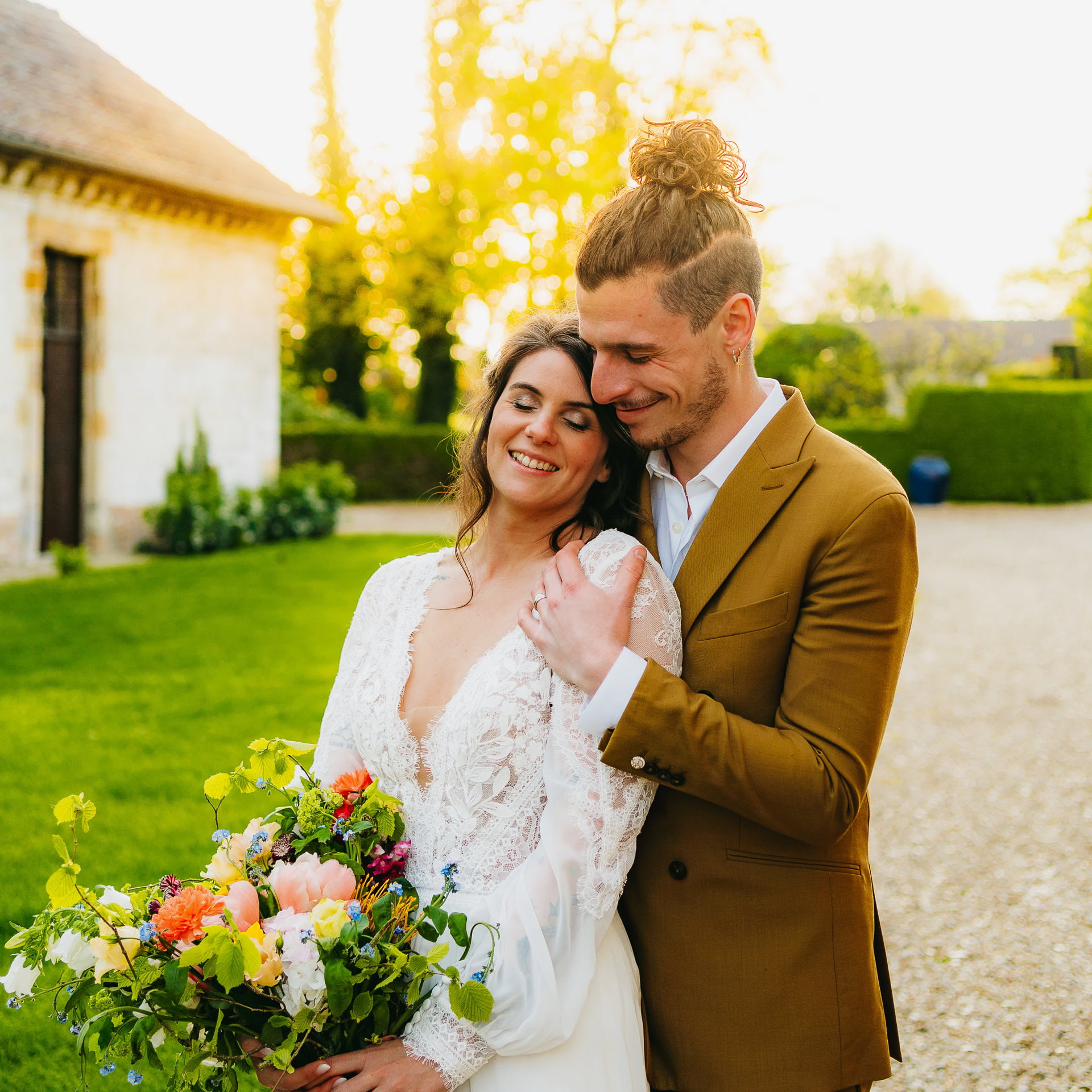 Un couple de jeunes mariés souriant et s'embrassant à l'extérieur, la mariée tenant un bouquet coloré, avec une douce lumière du soleil filtrant à travers les arbres en arrière-plan.