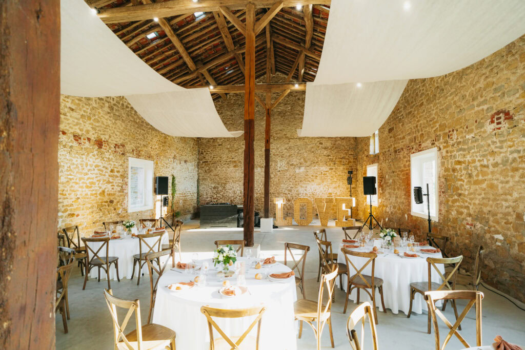 Un espace événementiel rustique avec des murs en pierre, des tables rondes dressées pour les repas et de grandes lettres formant le mot « LOVE », parfait pour une réception de mariage au charmant domaine de la Grange Lecomte dans les Ardennes. Un tissu drapé est élégamment suspendu au plafond.