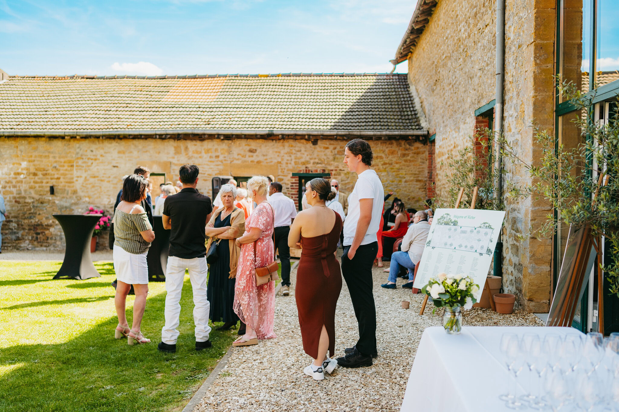 Un groupe de personnes se rassemble et discute dehors par une journée ensoleillée à côté d'un bâtiment rustique au Domaine de la Grange Lecomte. Une table avec quelques articles et un panneau d'affichage se trouvent à proximité, créant le cadre idéal pour un mariage dans les Ardennes.