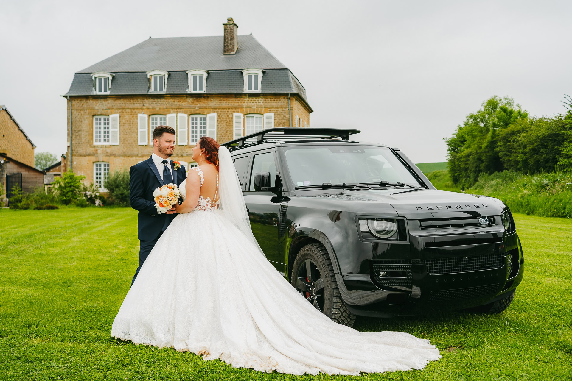 Une mariée et un marié se tiennent à côté d'un SUV noir sur une pelouse du prestigieux Domaine de la Grange Lecomte, avec une grande maison en briques en toile de fond, prêts à célébrer leur réception de mariage.