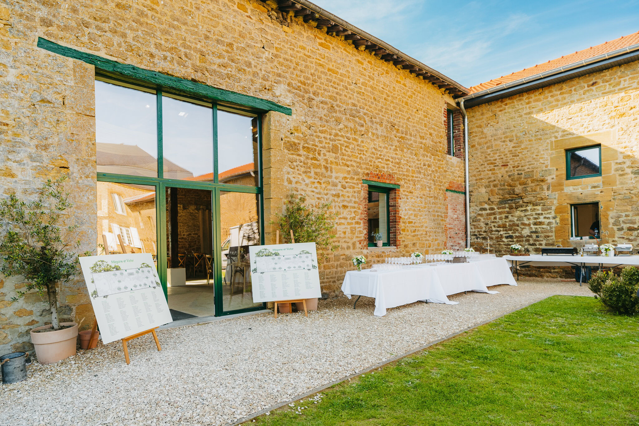 Aménagement de réception en plein air avec tables et chevalets blancs affichant les plans de table devant un bâtiment en pierre rustique dans le charmant Domaine de la Grange Lecomte, niché dans les pittoresques Ardennes. Parfait pour un mariage magique au milieu de la beauté de la nature.