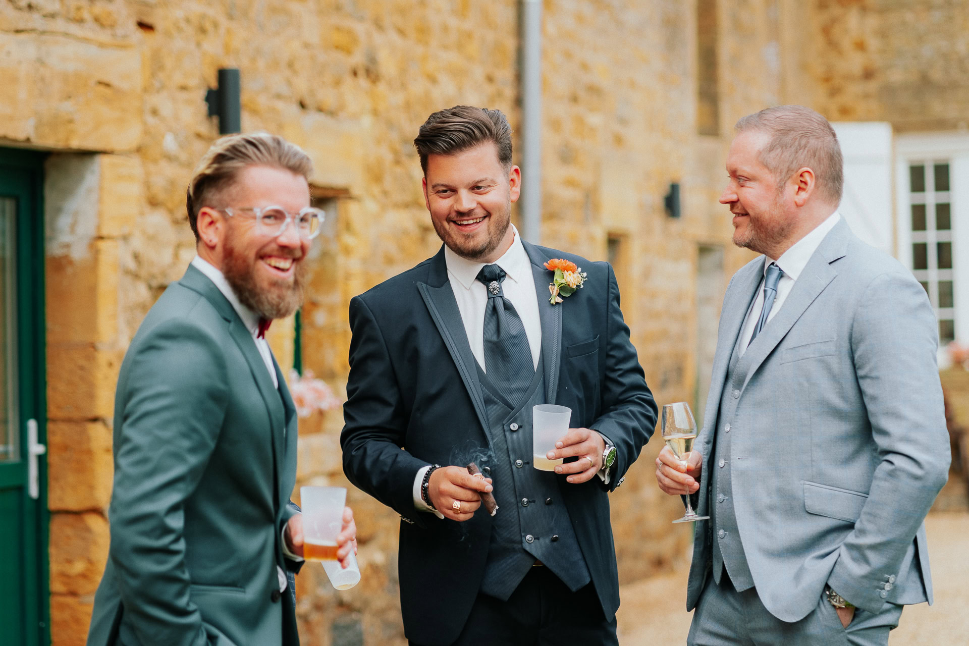 Trois hommes en costume se tiennent à l'extérieur, souriant et tenant des boissons lors d'une réception de mariage. L'un d'eux porte une boutonnière alors qu'ils se tiennent devant un bâtiment en pierre.