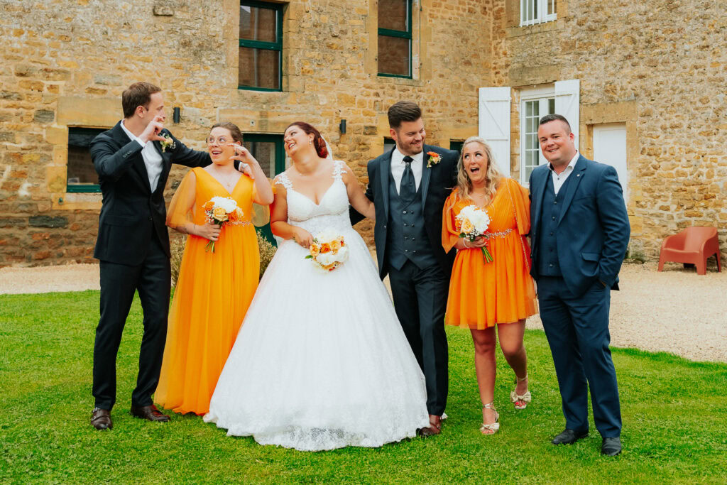 Un cortège nuptial de six personnes, dont trois en costume sombre et trois en robe orange vif, profite d'une conversation animée sur la pelouse luxuriante du Domaine de la Grange Lecomte. La mariée éblouit dans sa robe blanche sur le magnifique fond de pierre, créant le décor parfait pour cette réception de mariage ardennaise.