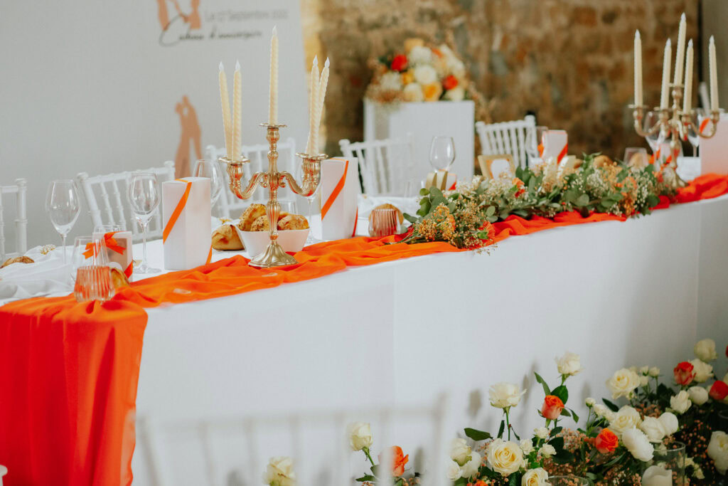 Table de mariage élégante au Domaine de la Grange Lecomte, avec chaises blanches, rideaux orange, bougies et centres de table fleuris. Assiettes, verres et couverts sont soigneusement disposés pour une occasion magique dans les Ardennes.