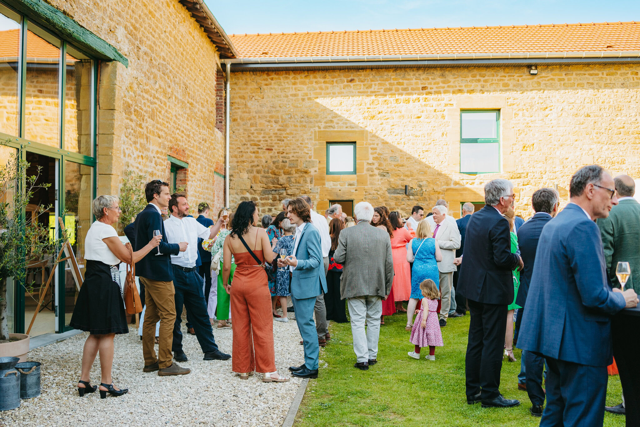 Lors d'un événement en journée, les invités se réunissent et discutent en plein air dans une charmante cour du Domaine de la Grange Lecomte. Entouré d'élégants bâtiments en pierre et d'espaces verts luxuriants, le cadre rappelle une réception de mariage enchanteresse dans les Ardennes.