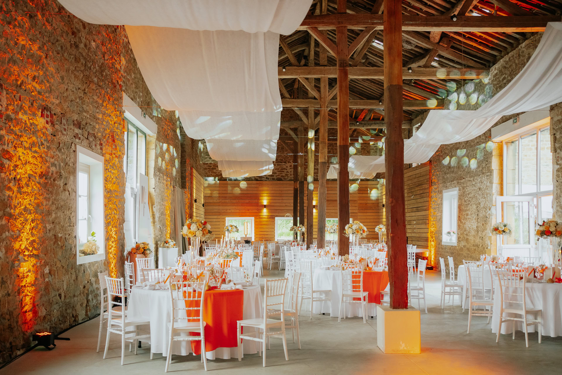 Organisez votre mariage rustique dans les magnifiques Ardennes, où une grange aux murs de pierre constitue le lieu de réception idéal. Profitez d'un plafond drapé de blanc et de tables rondes avec d'élégantes chaises blanches sous un éclairage chaleureux pour une soirée inoubliable.