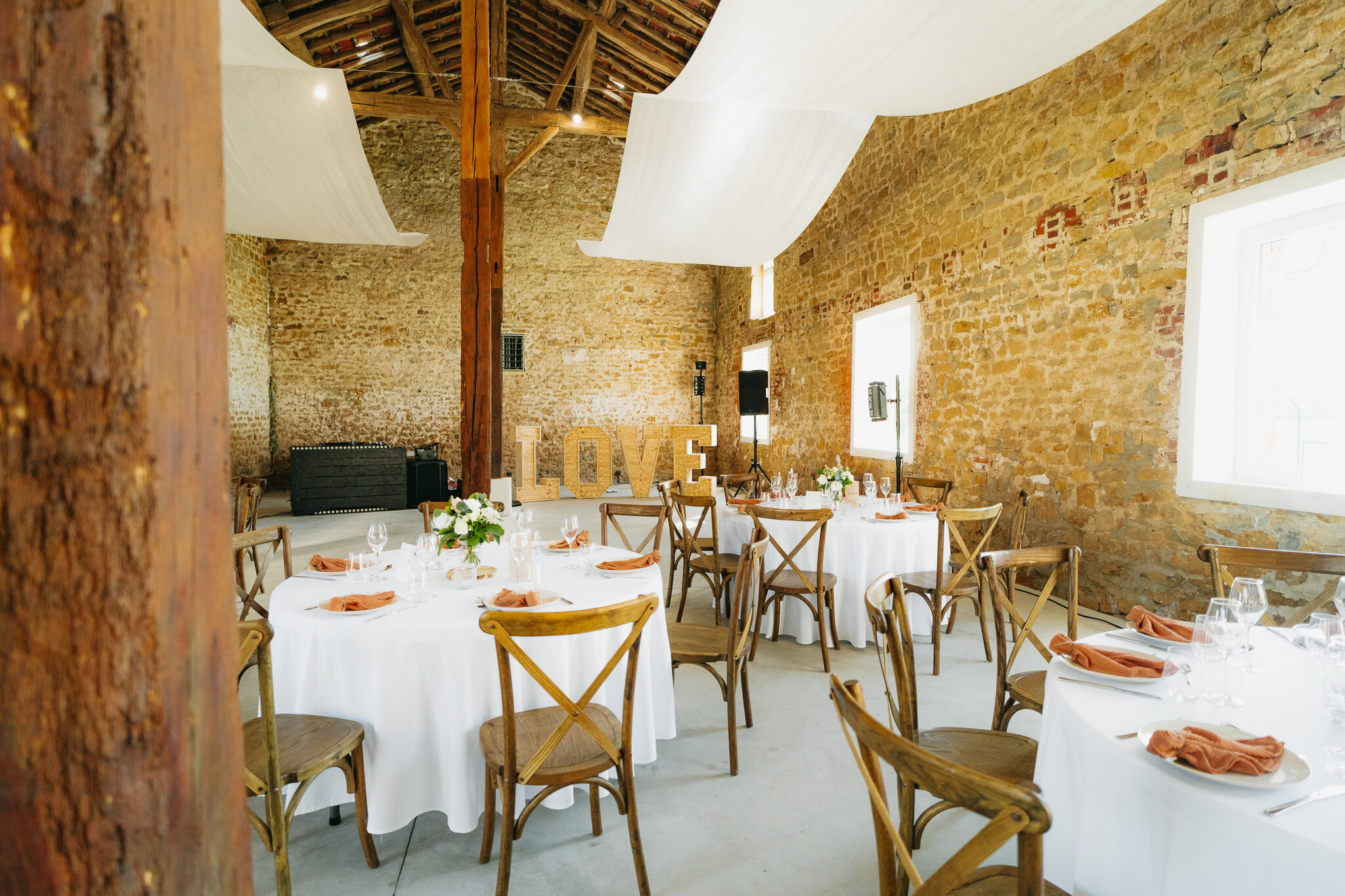 Une réception de mariage rustique dans les charmantes Ardennes, avec des tables rondes avec des nappes blanches, des chaises en bois et des centres de table floraux au Domaine de la Grange Lecomte. Un panneau « LOVE » orne un mur en pierre, tandis que des rideaux blancs pendent élégamment du plafond.