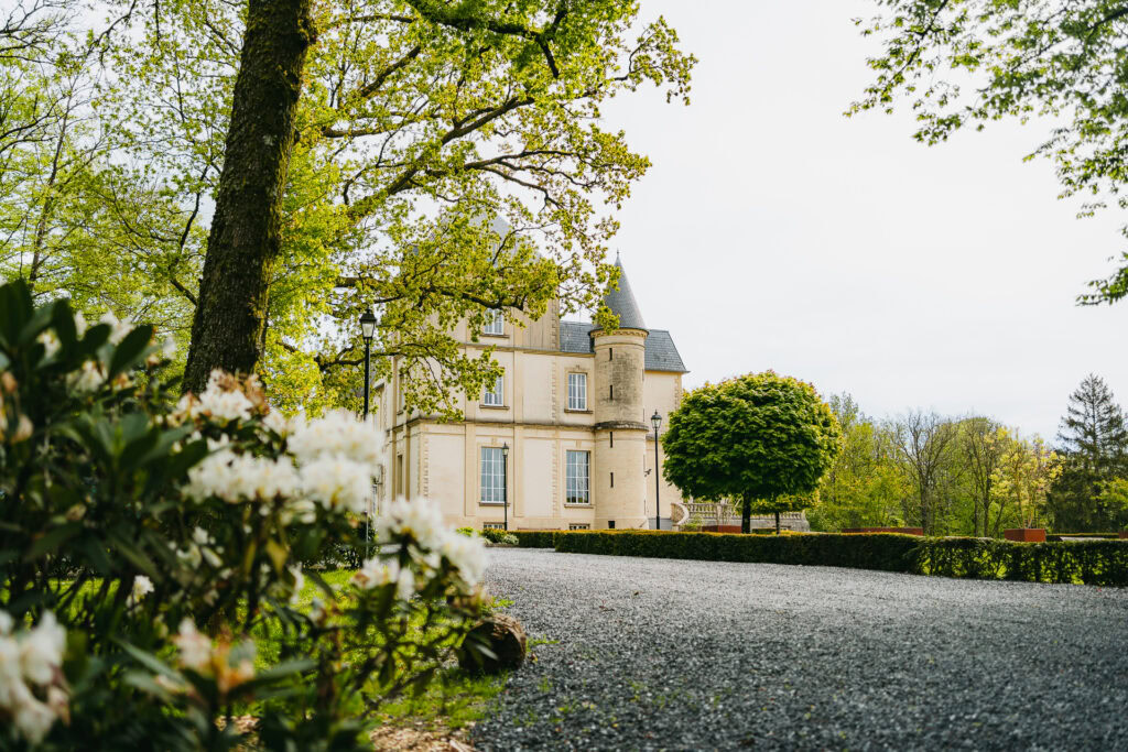 Le Château du Bois d'Arlon, bâtiment en pierre beige avec tourelle et fenêtres cintrées, se dresse derrière une allée de gravier, entouré d'arbres et d'arbustes fleuris dans un jardin paysager.