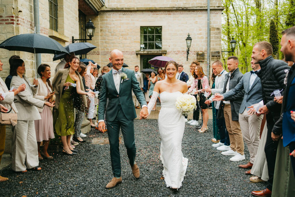 Une mariée et un marié marchent main dans la main dans une allée extérieure du Château du Bois d'Arlon, souriants, tandis que les invités des deux côtés applaudissent et célèbrent avec des parapluies et des confettis.