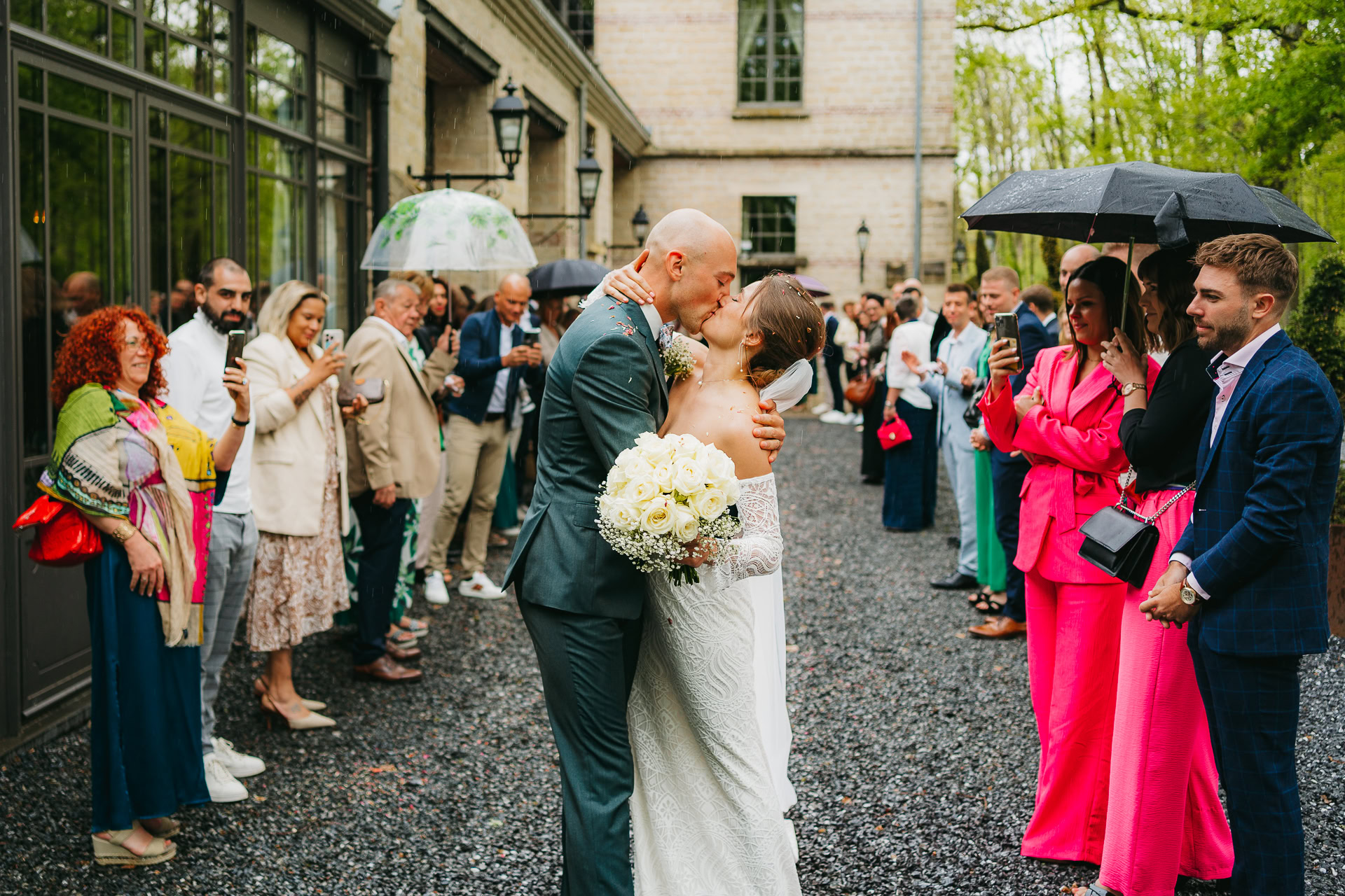 Un couple de mariés s'embrasse en plein air au Château du Bois d'Arlon après leur cérémonie de mariage sous le regard des invités, certains tenant des parapluies un jour de pluie.