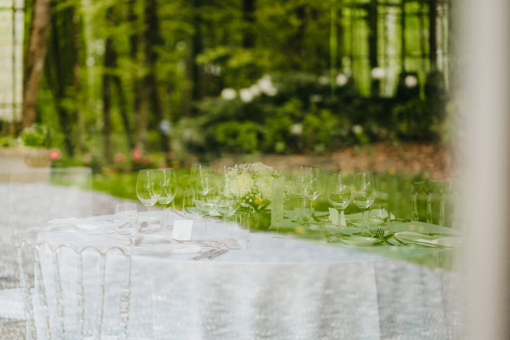 Une table ronde dressée avec de la verrerie, des assiettes et un centre de table floral est visible à travers une fenêtre du Château du Bois d'Arlon, avec des reflets de l'espace extérieur luxuriant et boisé.