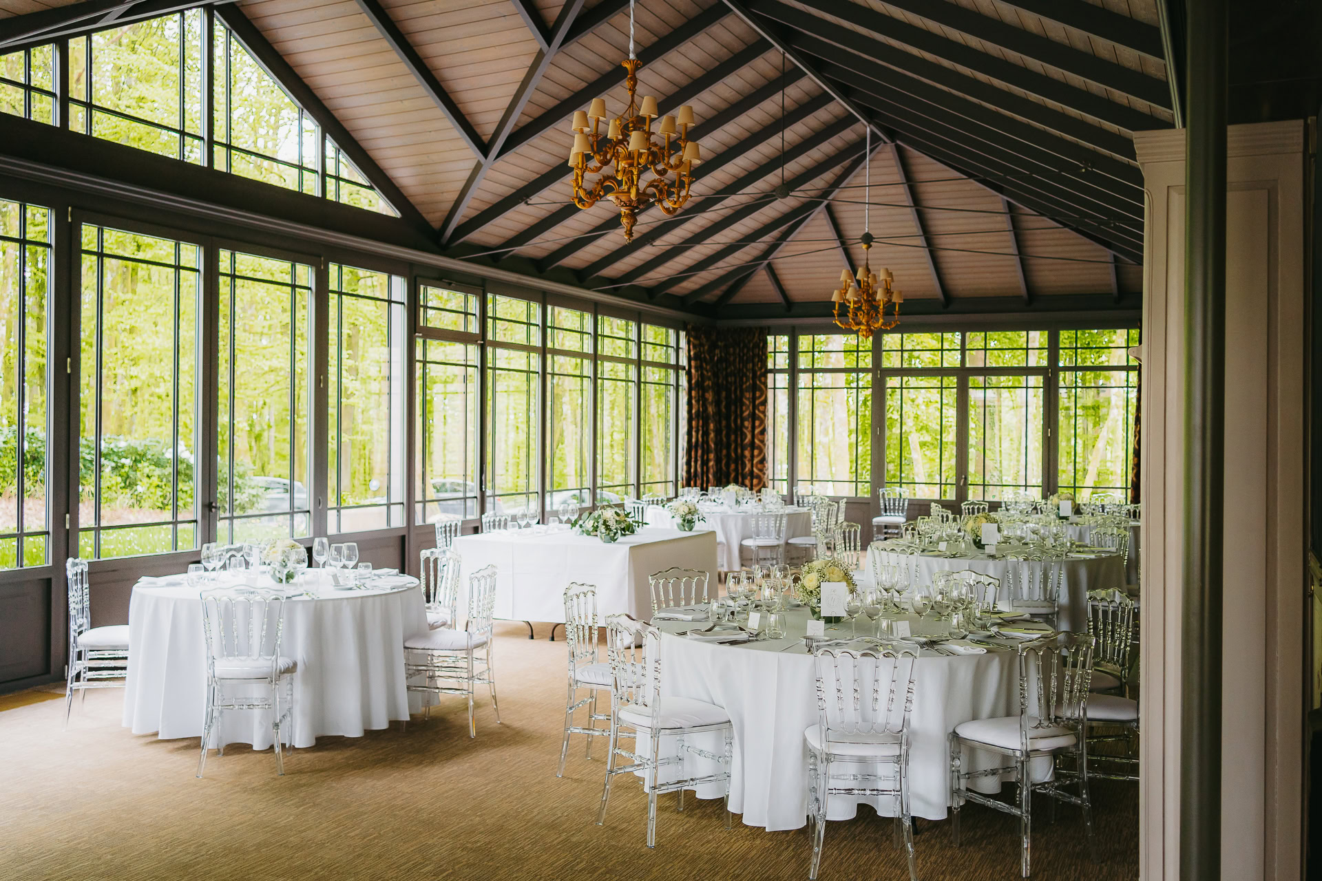 Une salle à manger lumineuse et élégante du Château du Bois d'Arlon dispose de grandes fenêtres, de tables rondes avec des nappes blanches, des chaises transparentes et des lustres suspendus à un plafond voûté.