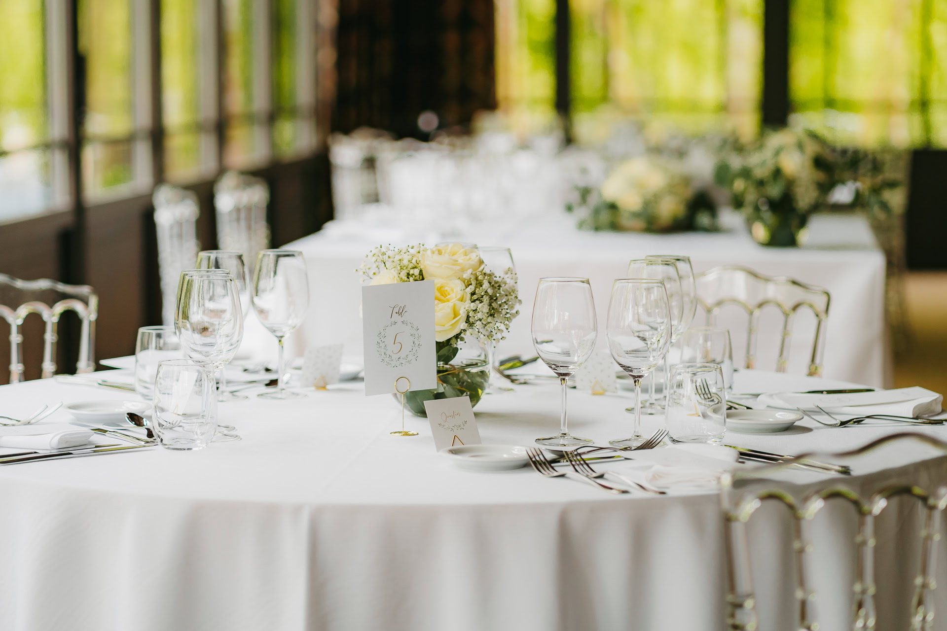 Une table de banquet ronde au Château du Bois d'Arlon est dressée avec du linge blanc, de la verrerie, des assiettes, de l'argenterie et une pièce maîtresse florale ; une carte de numéro de table est affichée au centre.