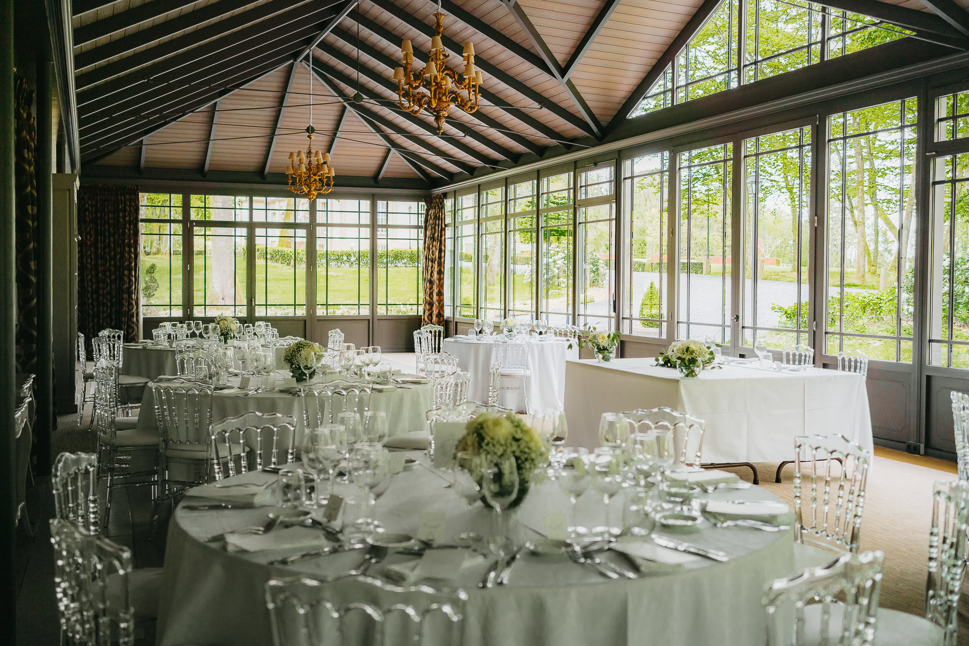 Un espace événementiel lumineux et vitré au Château du Bois d'Arlon aménagé pour un repas formel avec des tables rondes, des nappes blanches, des chaises transparentes et des centres de table floraux.