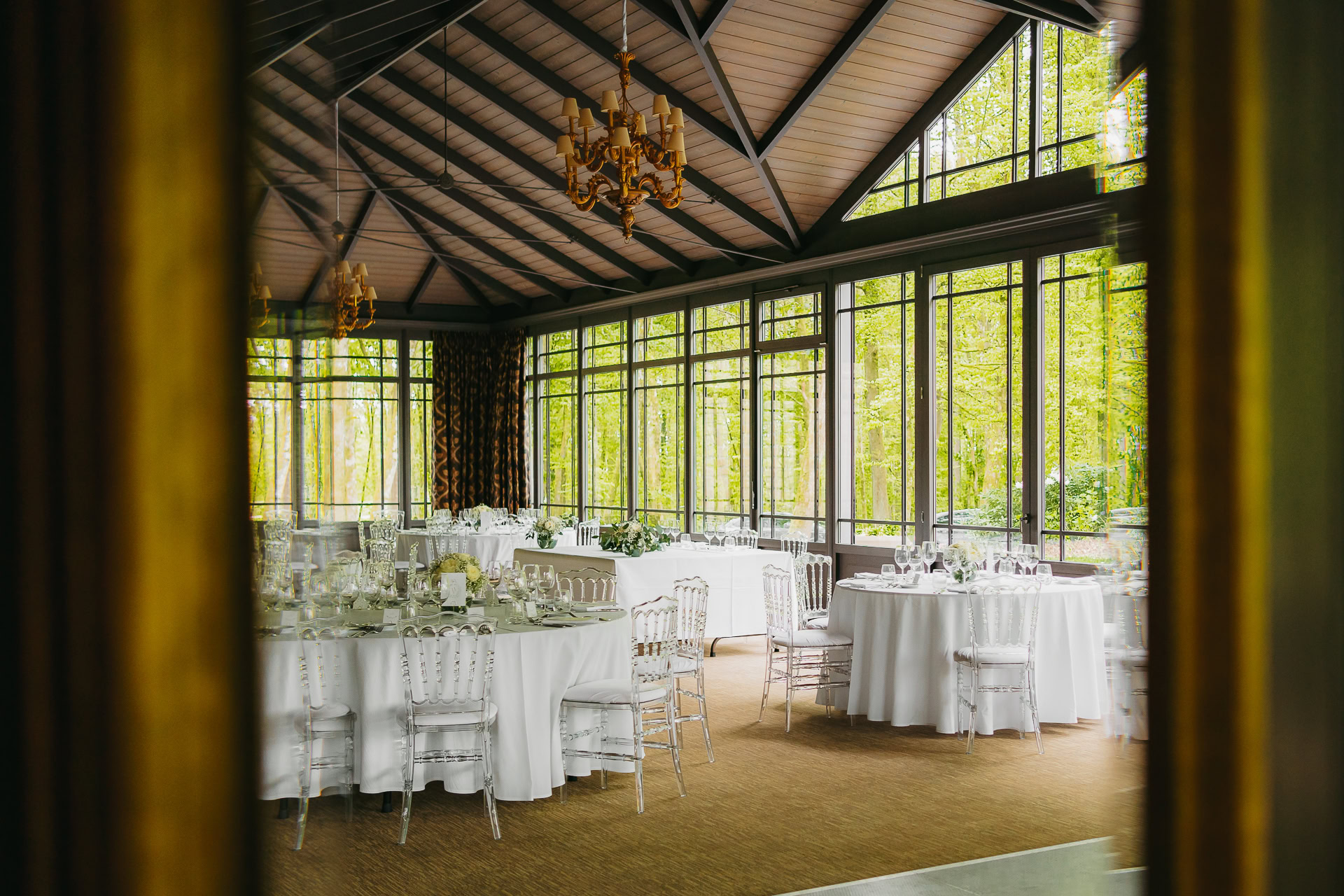 Une salle de banquet au Château du Bois d'Arlon avec de grandes fenêtres, des tables rondes drapées de nappes blanches, des chaises claires et des couverts élégants ; une verdure luxuriante visible à l'extérieur.
