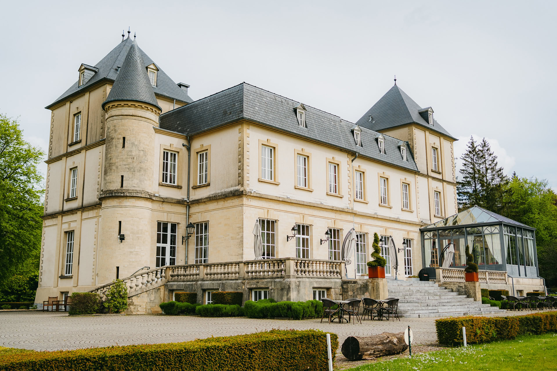 Grand château en pierre beige doté de multiples fenêtres, de tourelles et d'une véranda vitrée, le Château du Bois d'Arlon est entouré de verdure et d'une cour pavée.