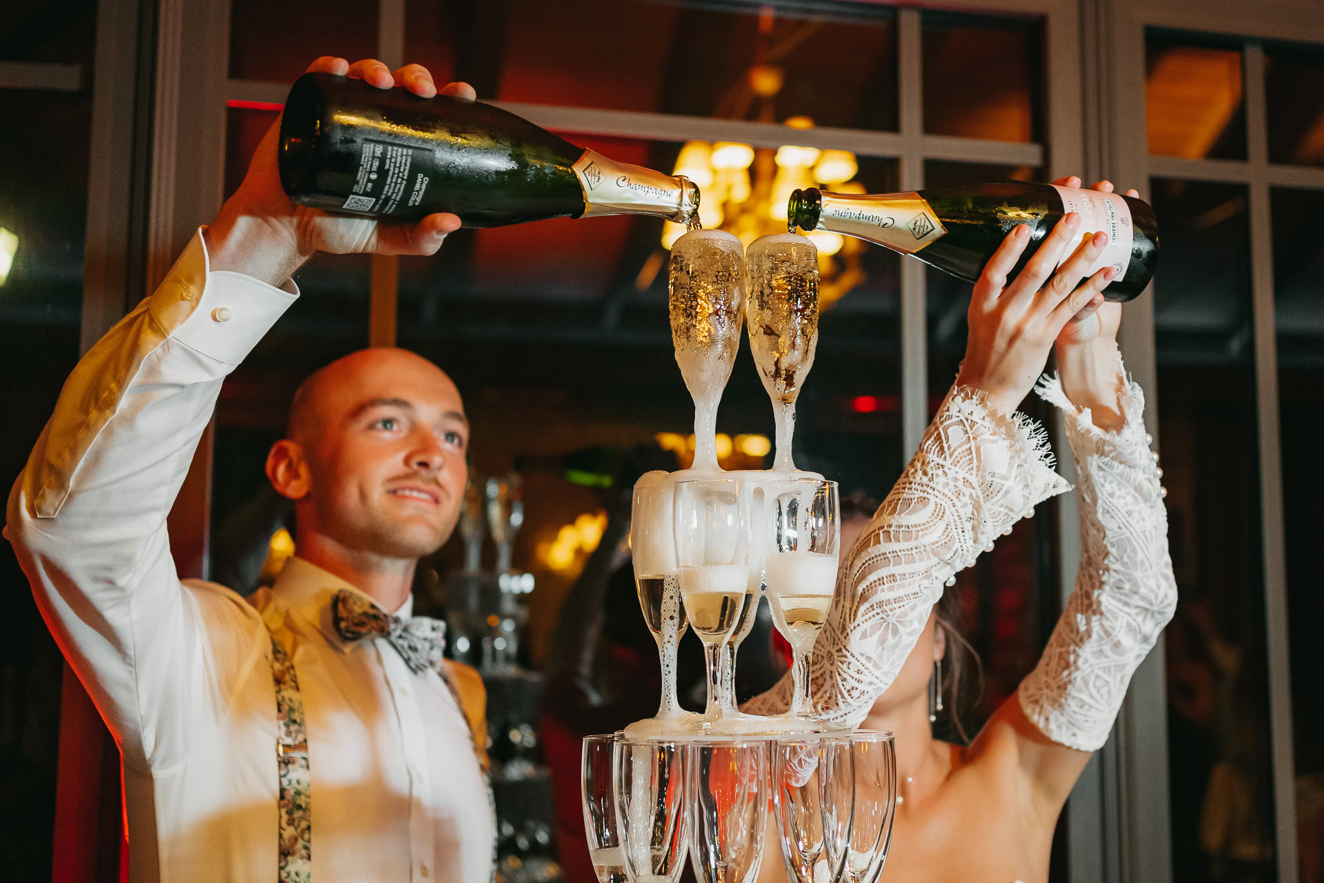 Lors d'un événement au Château du Bois d'Arlon, un homme et une femme versent du champagne dans une tour de verres remplis, tous deux tenant des bouteilles au-dessus de la pyramide en cascade.