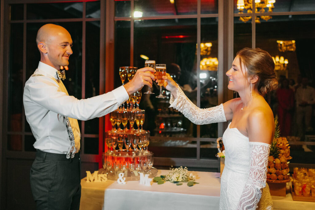 Un couple de mariés trinquent lors de leur réception de mariage au Château du Bois d'Arlon, debout près d'une tour de champagne et d'une table joliment décorée.