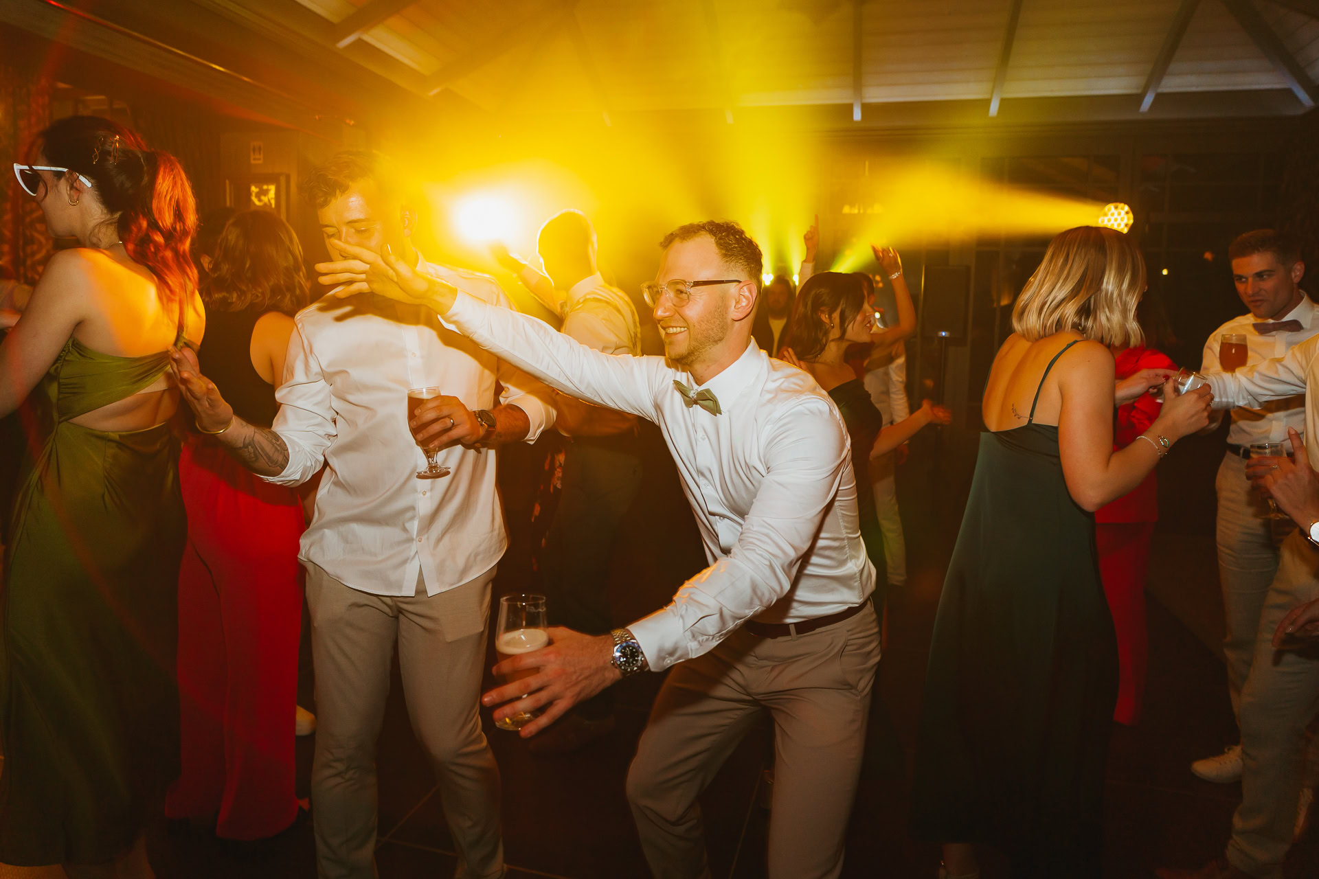Des gens dansent et socialisent lors d'une fête dans la chaude lueur jaune du Château du Bois d'Arlon, tandis qu'un homme devant tend la main avec un verre à la main.