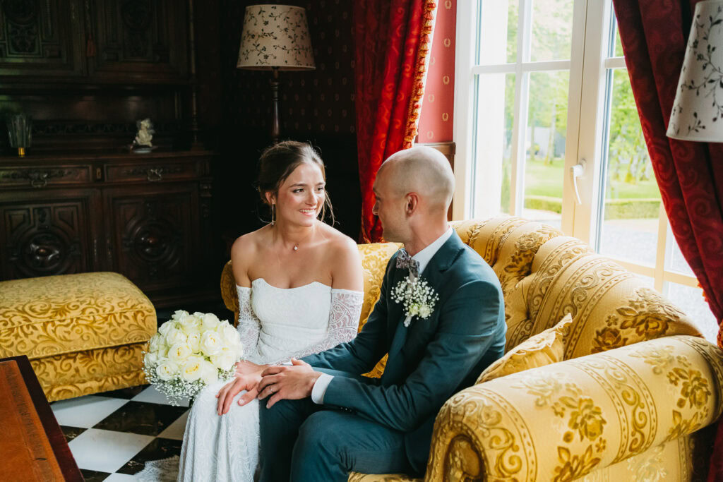 Une mariée en robe blanche et un marié en costume sont assis ensemble sur un canapé à motifs jaunes au Château du Bois d'Arlon, se tenant la main et se souriant.