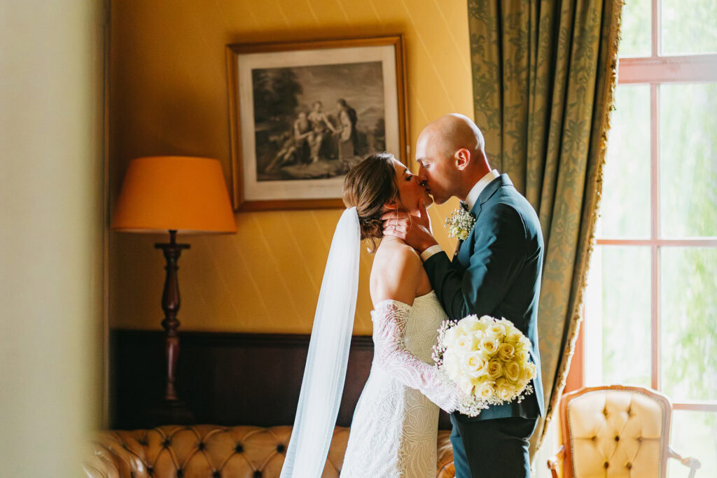 Une mariée et un marié partagent un baiser à l'intérieur près d'une fenêtre du Château du Bois d'Arlon, la mariée tenant un bouquet de roses blanches.