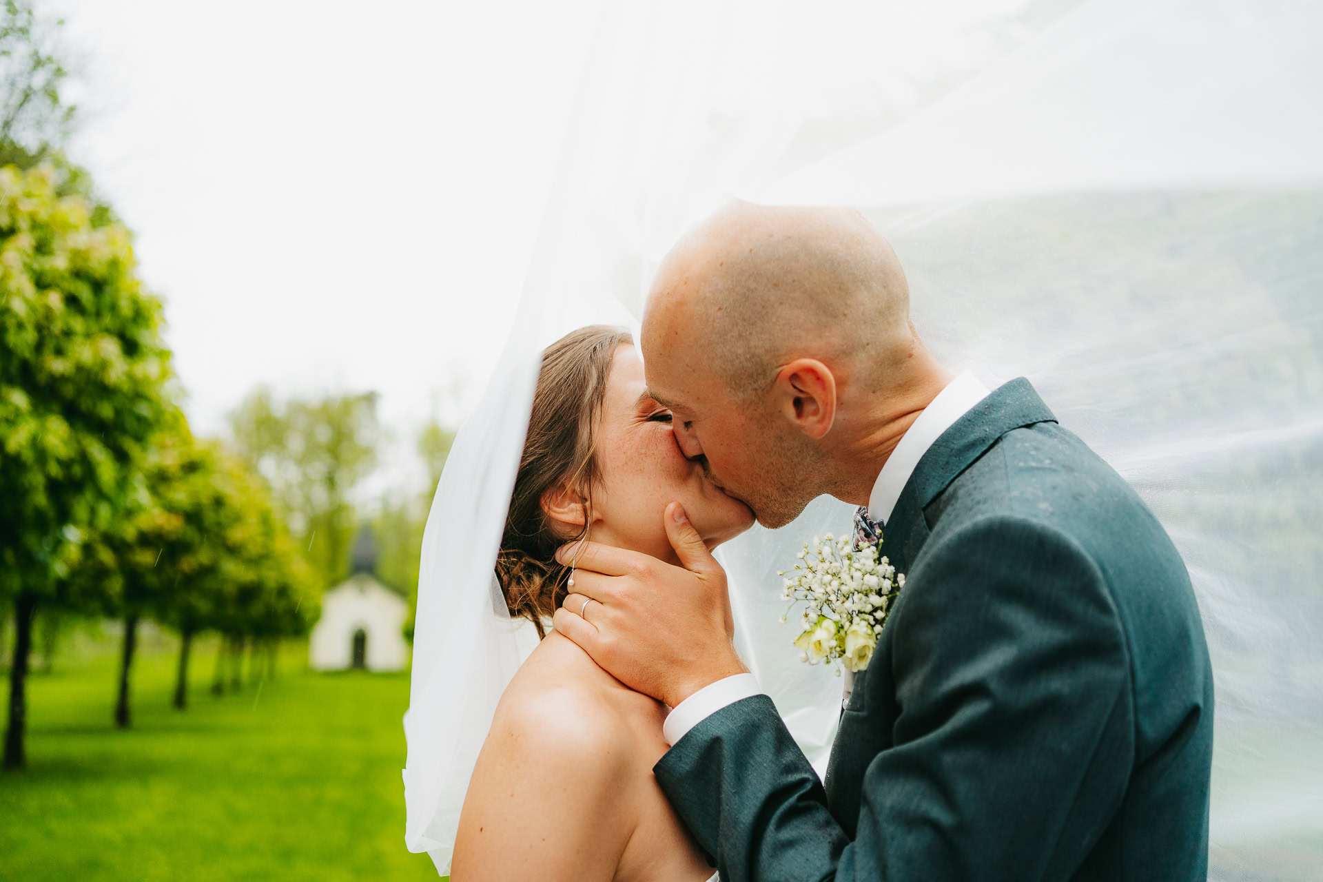 Un couple de mariés s'embrasse en plein air au Château du Bois d'Arlon. Le marié tient délicatement le visage de la mariée, son voile flottant derrière eux. Des arbres et un petit bâtiment complètent cette scène romantique en arrière-plan.